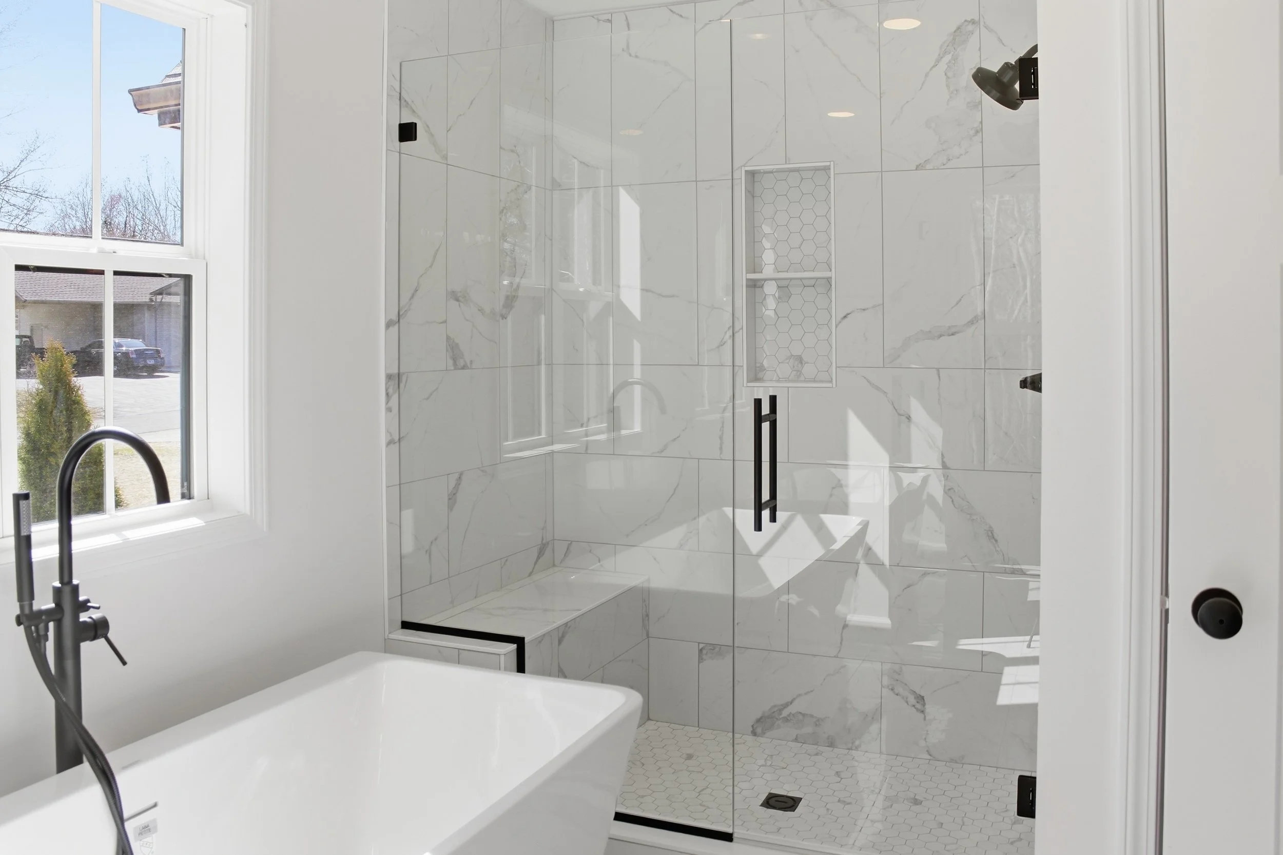 Modern bathroom with a large glass-enclosed shower featuring white marble tiles with gray veining, a built-in niche with hexagonal tile pattern, a white bathtub with a black faucet, and a window allowing natural light.