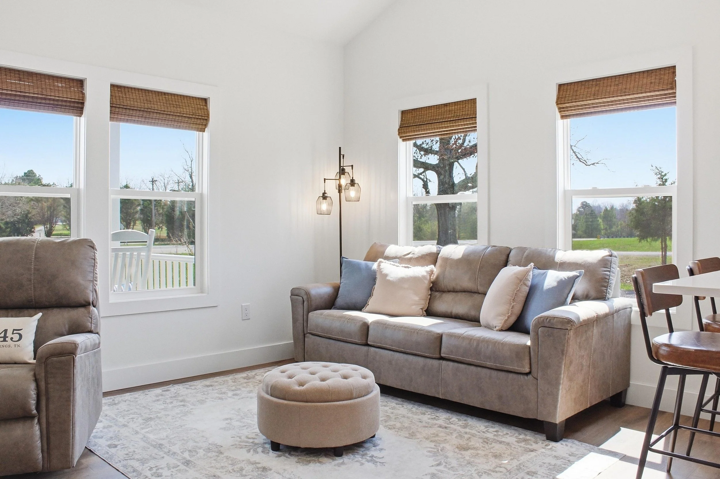 Living room with beige sofas, large windows with bamboo blinds, a standing floor lamp with exposed bulbs, a small round ottoman, and a patterned area rug.