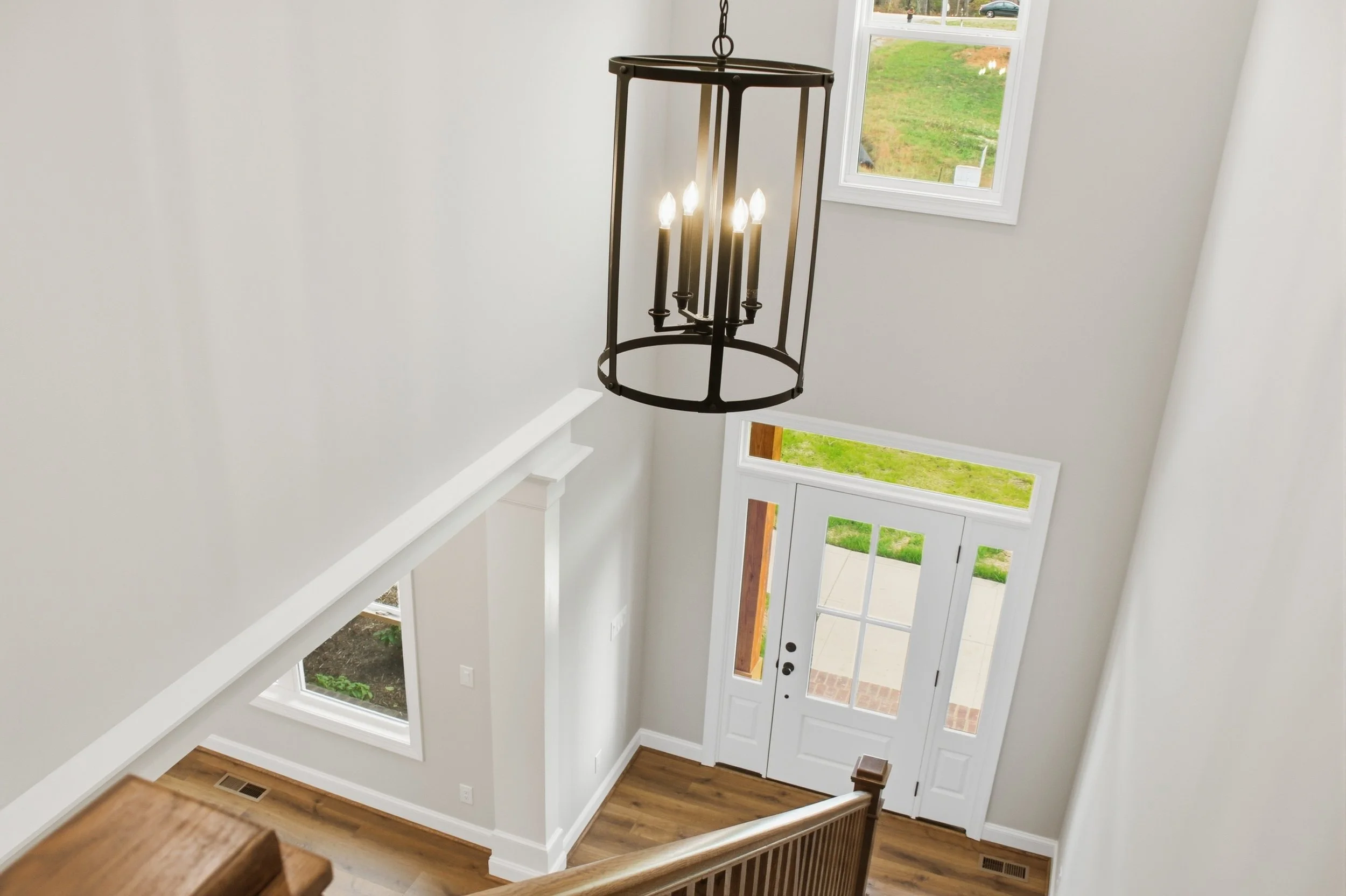 Entryway of a house with white walls, a front door with glass panels, hardwood flooring, and a black chandelier hanging from the ceiling.