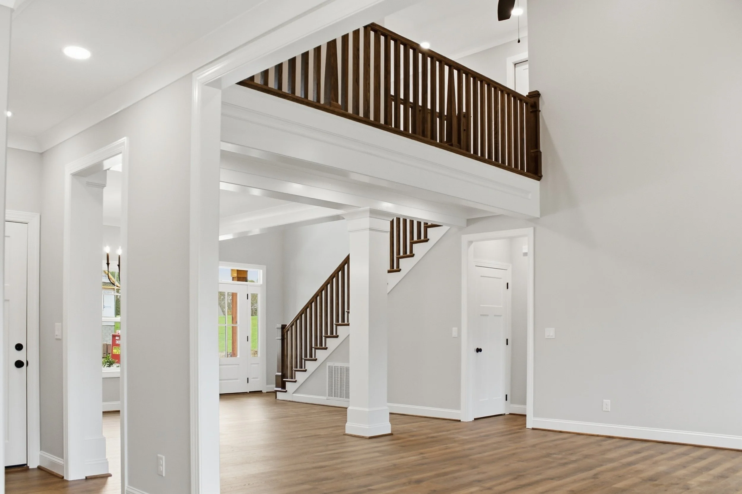 Interior view of a modern home with a staircase featuring wooden railing, white walls, and hardwood flooring. Multiple doorways and windows are visible, with natural light illuminating the space.