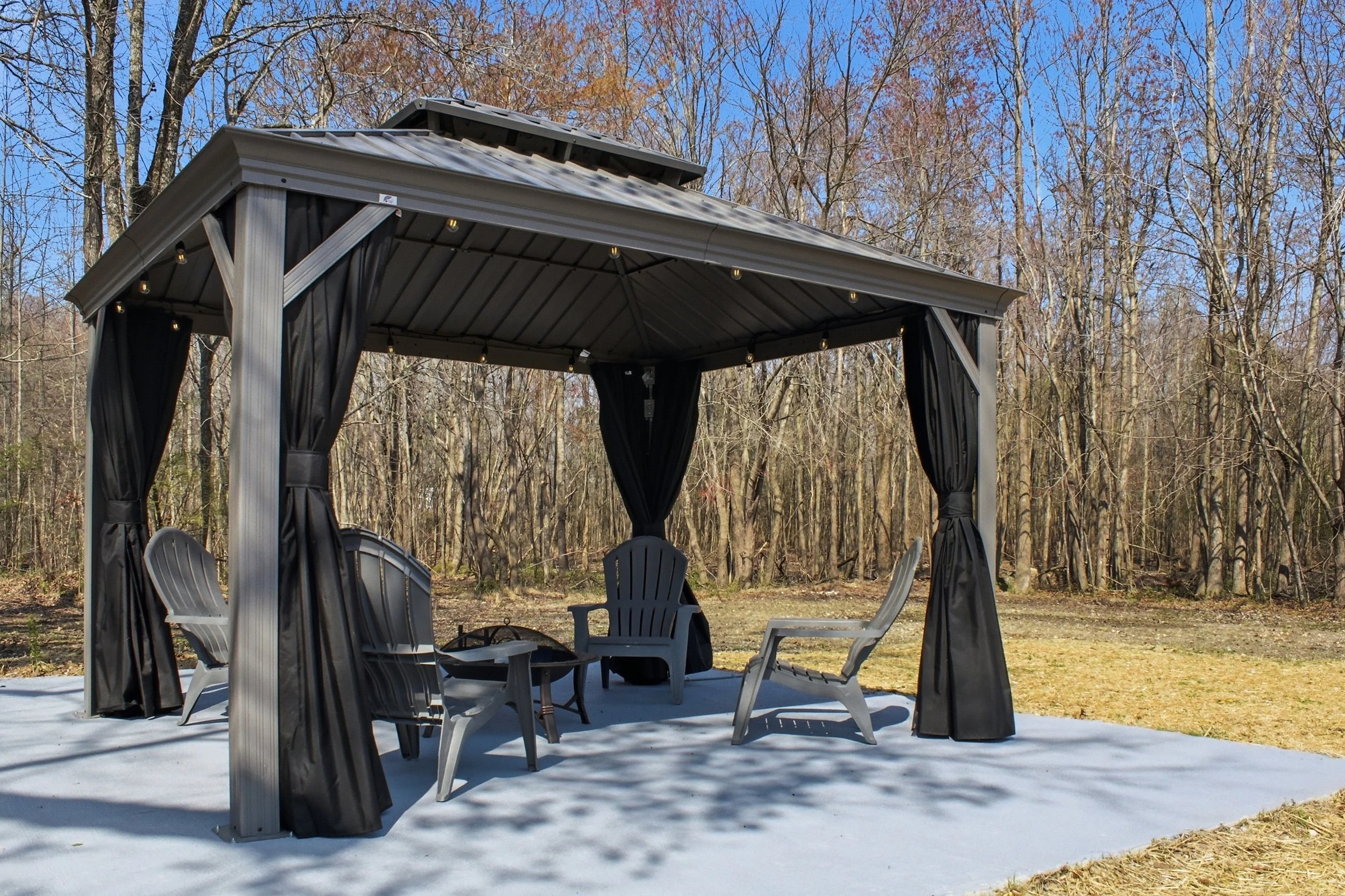 Outdoor pavilion with a metal roof, black curtains, and plastic chairs, surrounded by leafless trees and a blue sky.