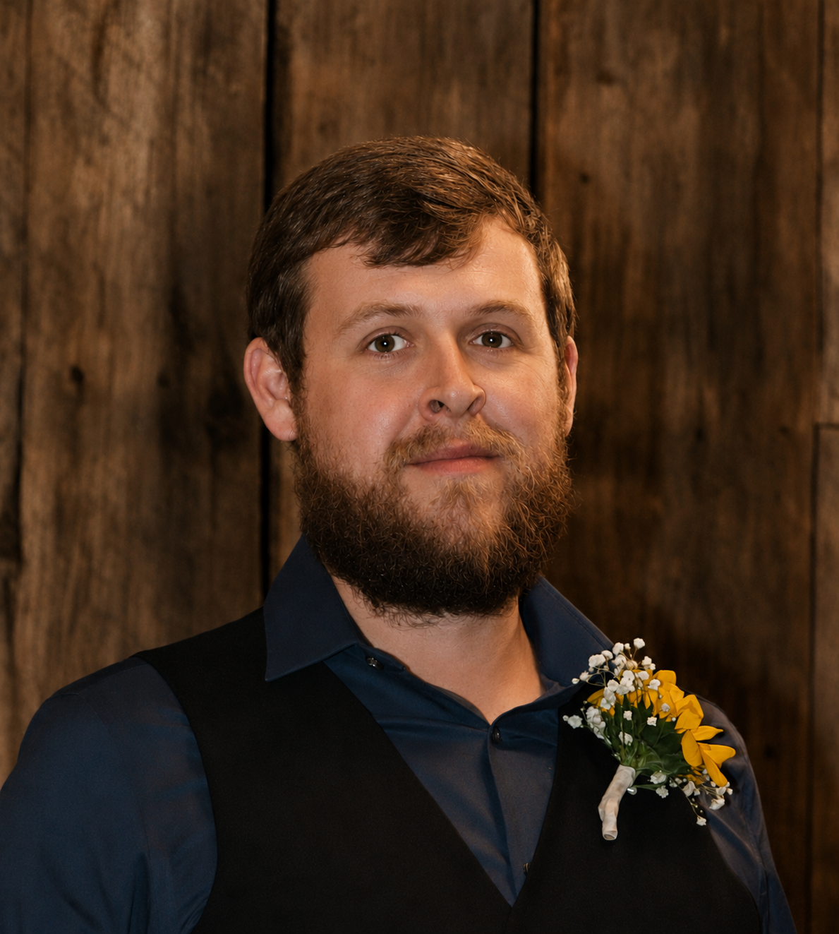 A man with a beard and brown hair wearing a dark blue shirt and a black vest, standing in front of a wooden wall, with a small boutonniere of yellow and white flowers on his vest.