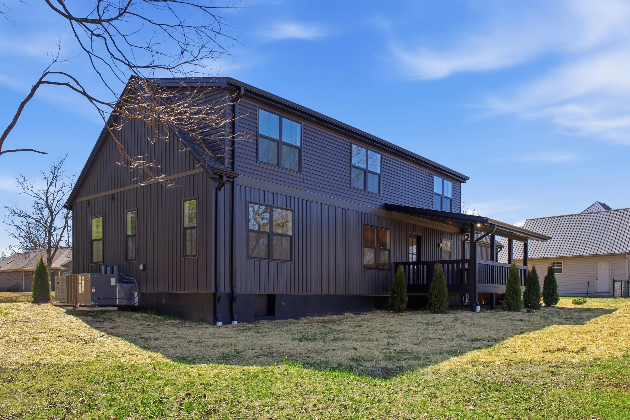 Modern multi-story house with dark siding, large windows, and a backyard with small trees, under a blue sky with some faint clouds.