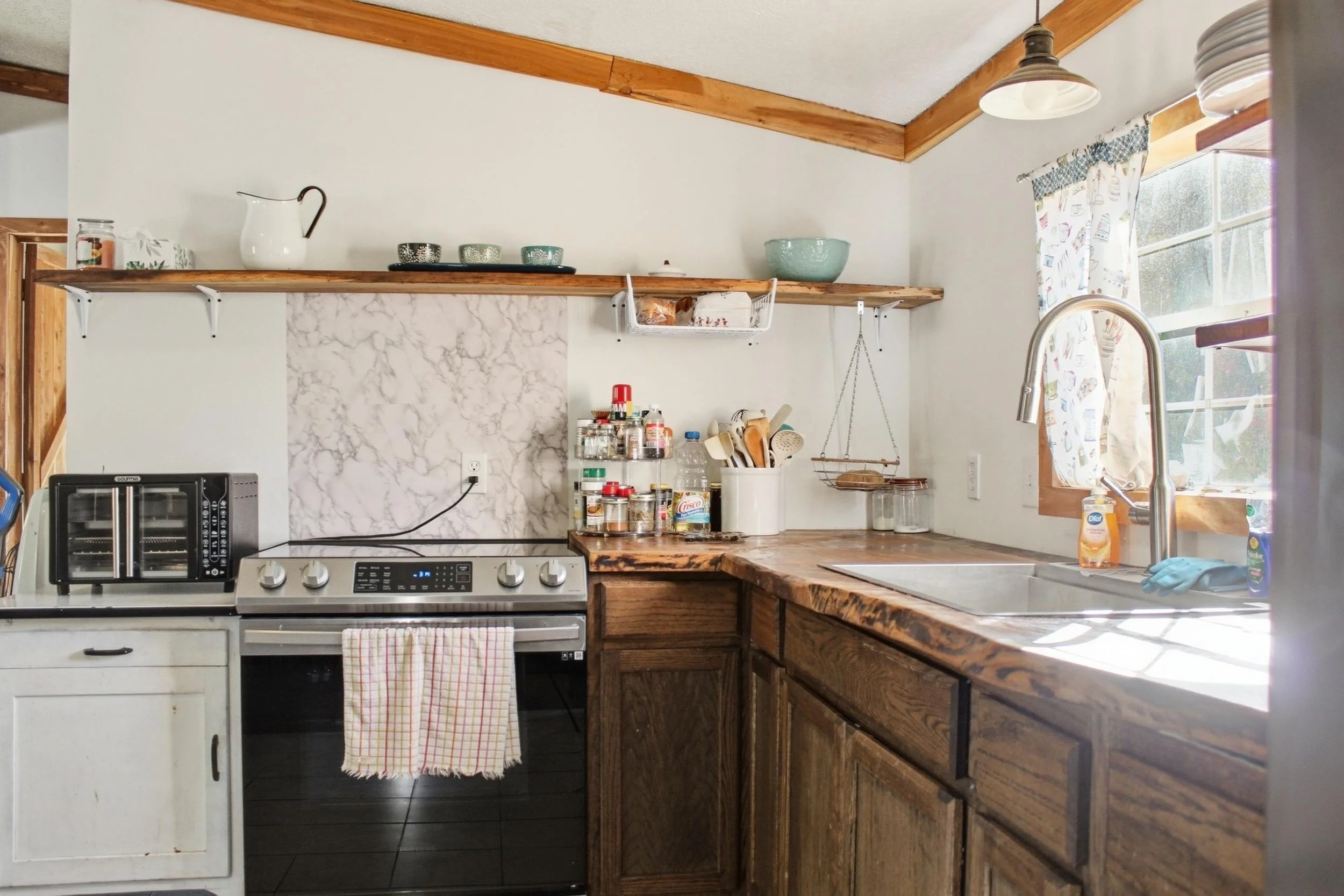 A kitchen with wooden countertops, white cabinets, a stove, and a sink near a window with patterned curtains. Open shelves hold dishes and jars, and various kitchen items are on the counter.
