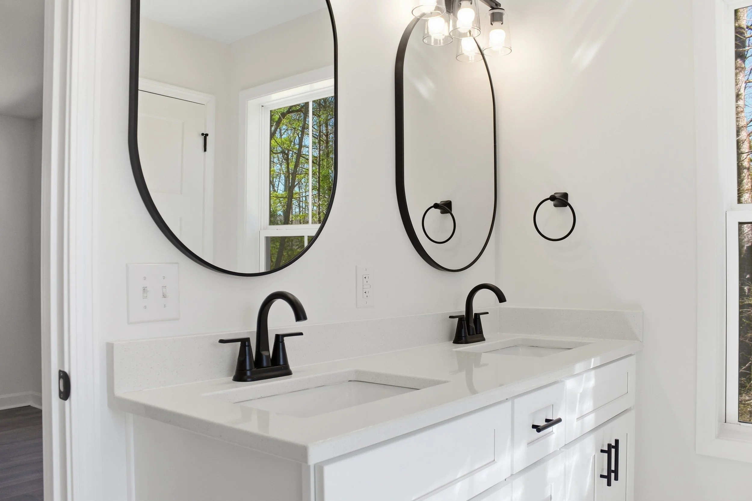 Modern bathroom with double vanity, black faucets, oval mirrors, pendant light fixtures, and windows showing trees outside.