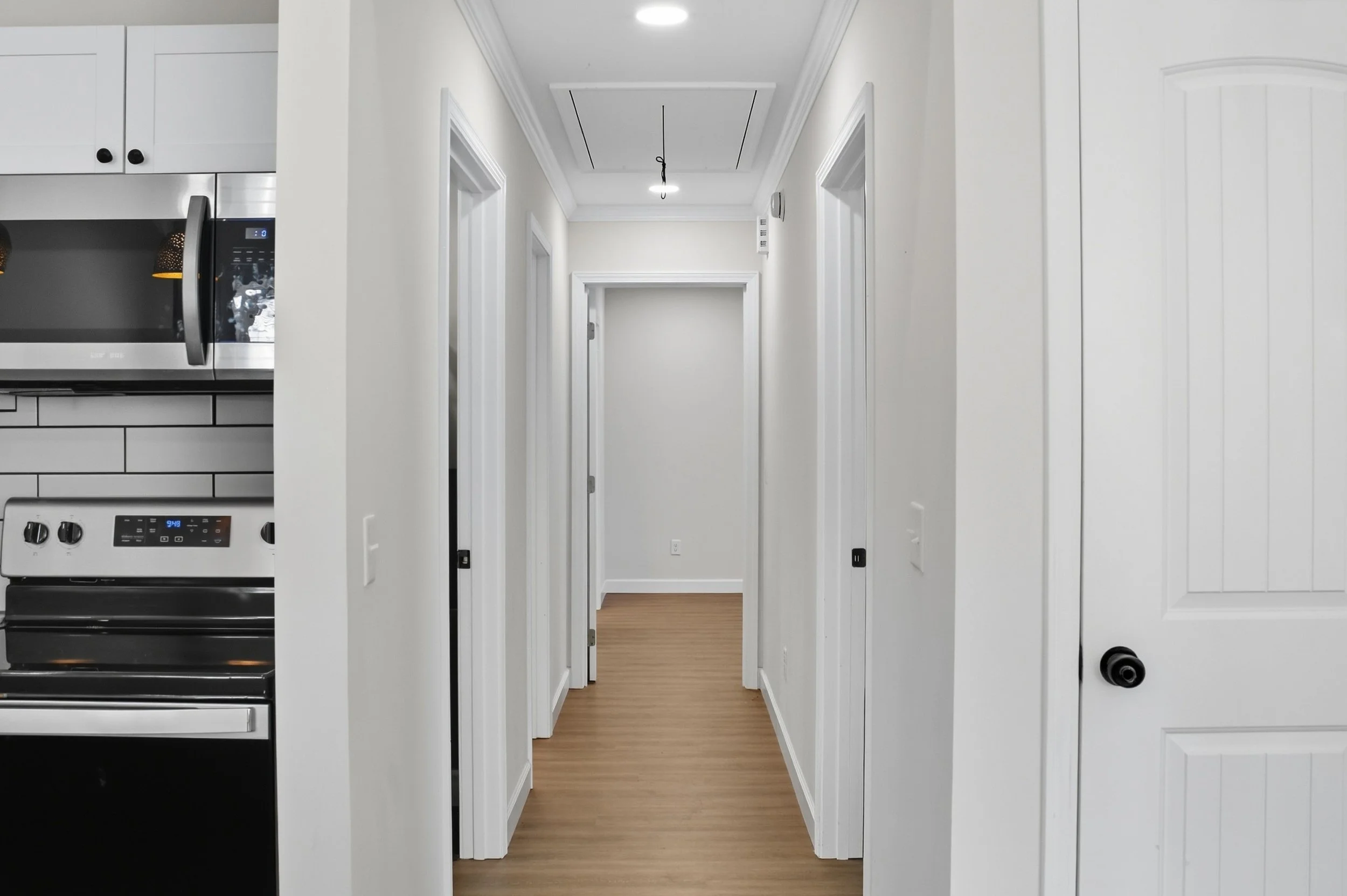 View of a hallway with white walls and doors, brown wood flooring, and a ceiling with recessed lighting, leading to another room in a modern home.