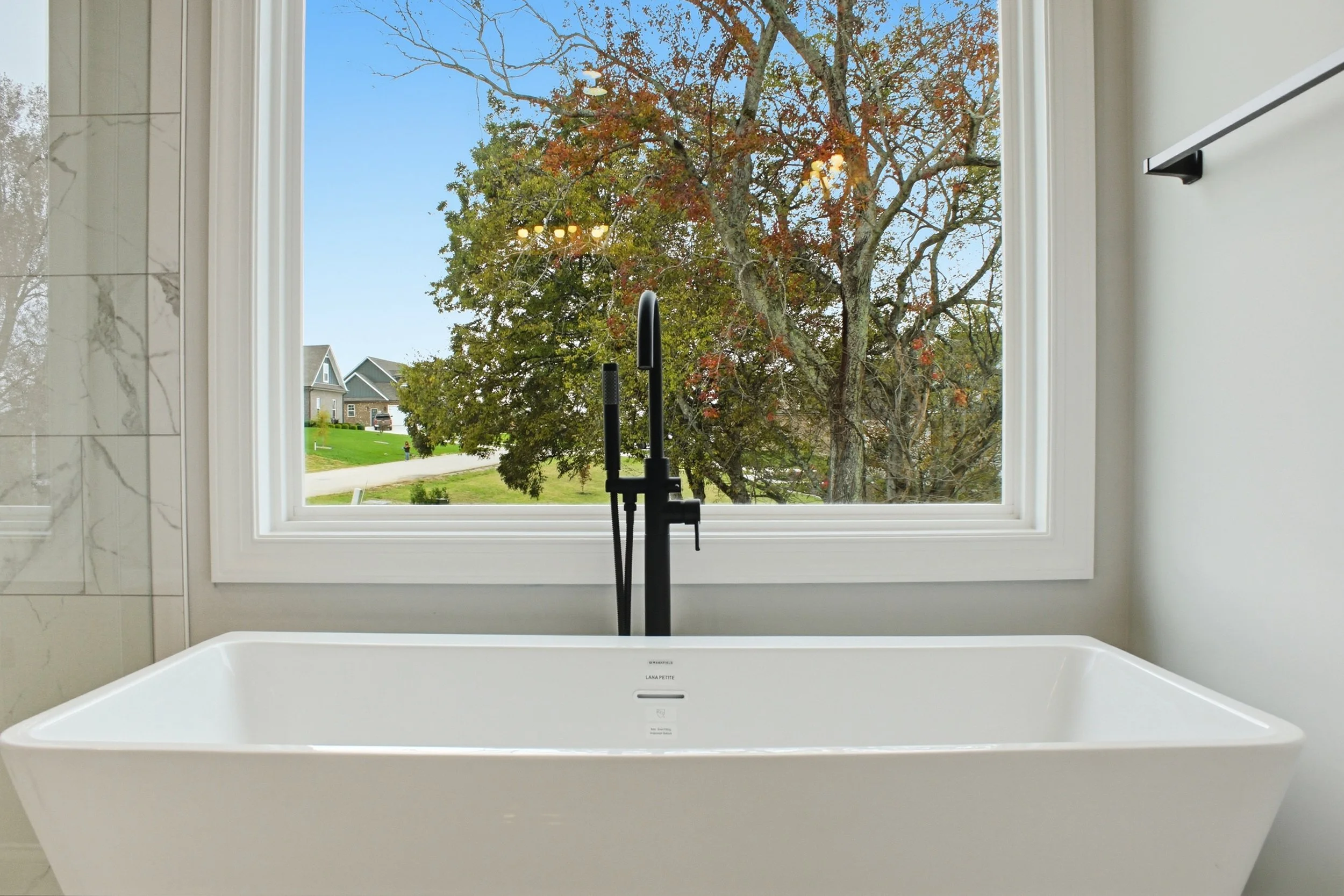 A modern walk-in bathtub with a large window overlooking a tree with autumn leaves and part of a neighborhood outside.