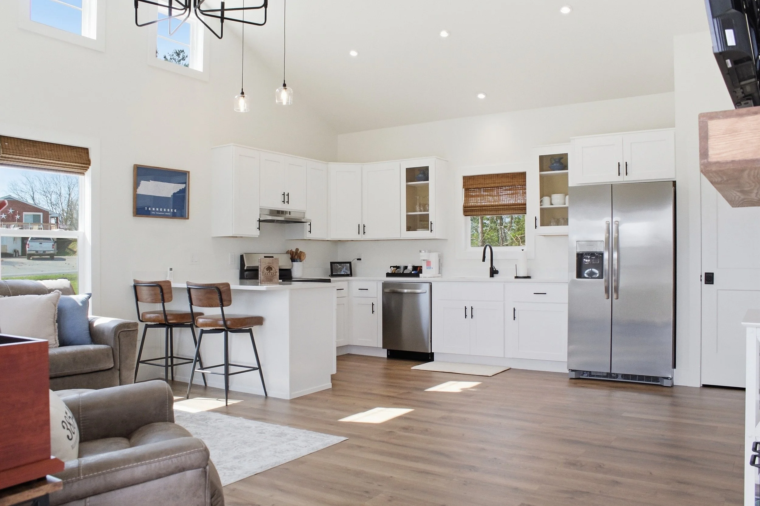 Modern kitchen with white cabinets, stainless steel refrigerator, island with barstools, wooden flooring, and windows with bamboo blinds.