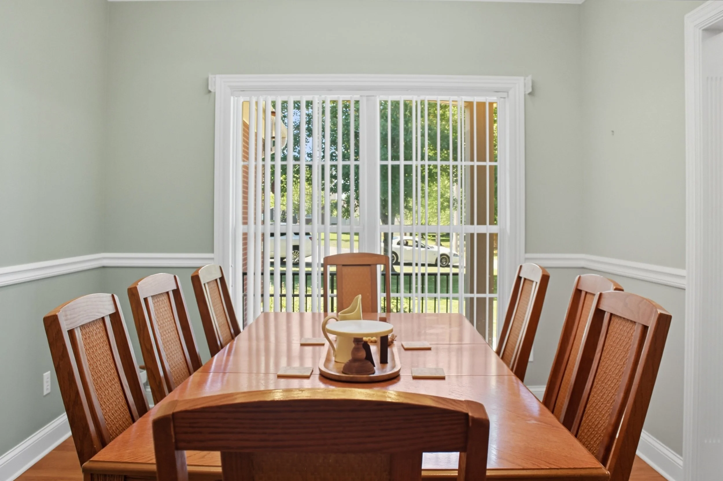 Dining room with wooden table and chairs, large window with vertical blinds showing trees and parked cars outside.