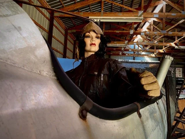 A mannequin dressed as a pilot in a leather jacket and vintage helmet inside an airplane fuselage in a hangar.
