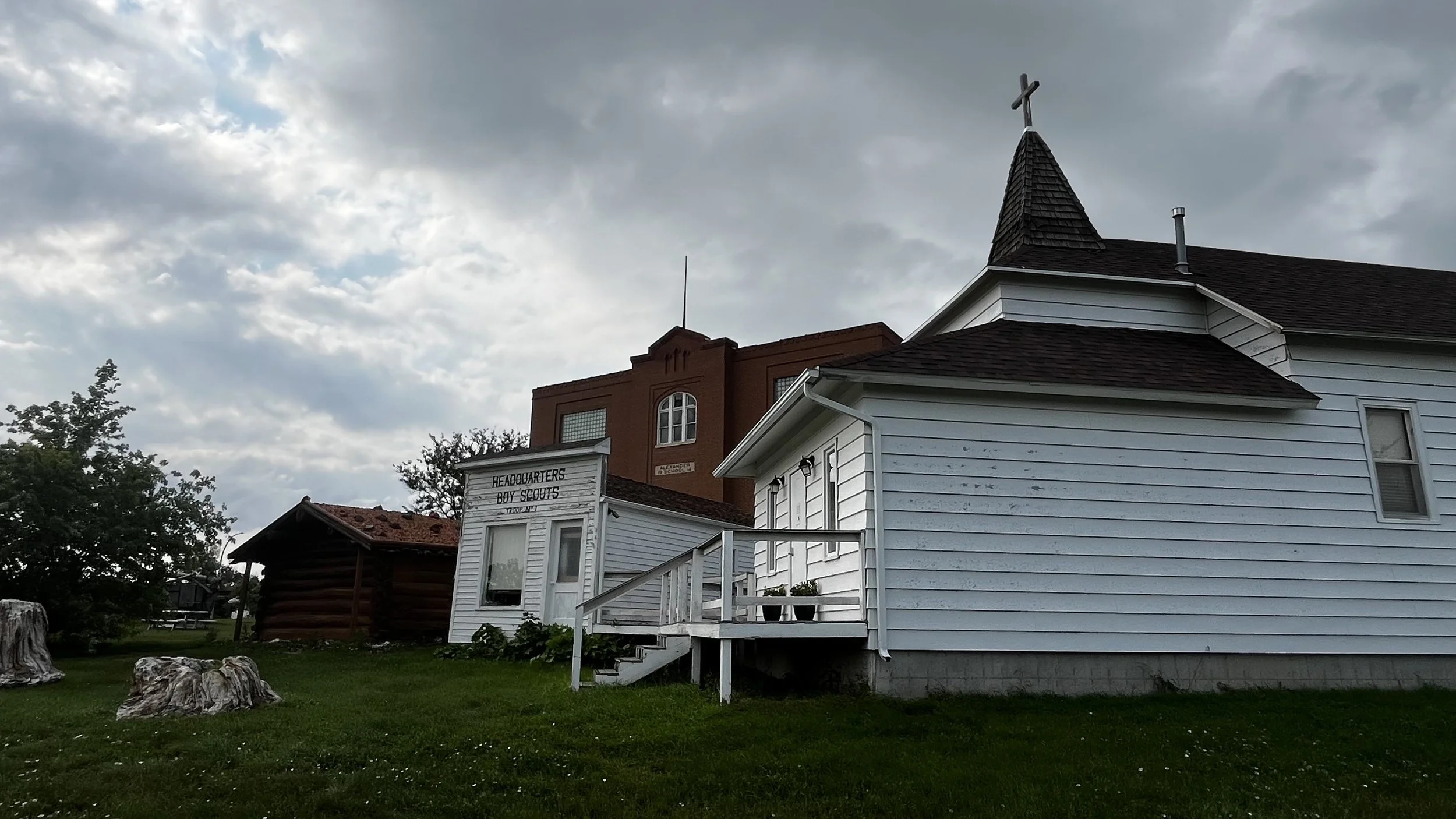 A church-like building with white wooden siding and a small steeple with a cross on top. Behind it is a taller red brick building. The sky is cloudy, and there are some large rocks on the grass in the foreground. A sign reads "Headquarters Boy Scouts."