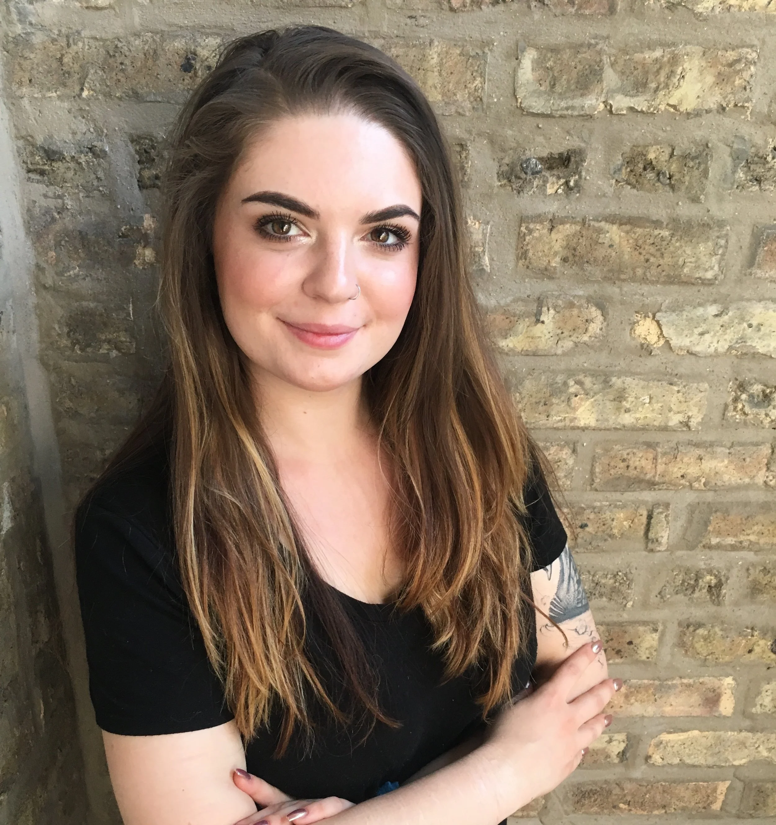 A young woman with long brown hair, light complexion, and hazel eyes, smiling softly, standing in front of a brick wall, wearing a black top.