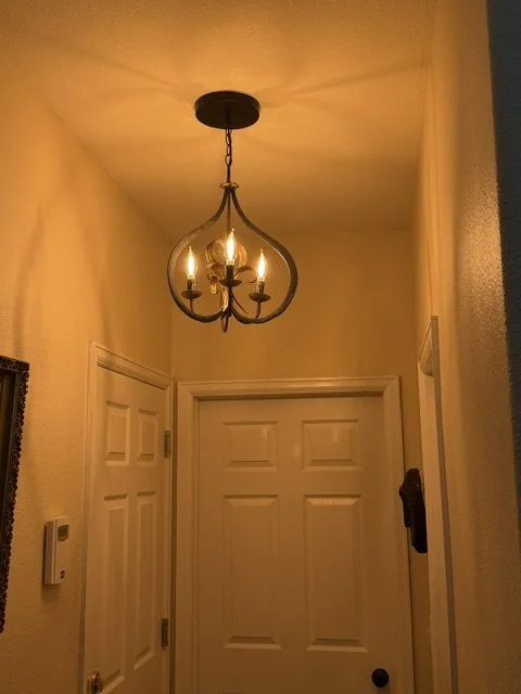 A hallway ceiling with a black metal chandelier with four candle-shaped lights, illuminating the space. There are three white doors, one on the left and two at the end, with a thermostat on the left wall.