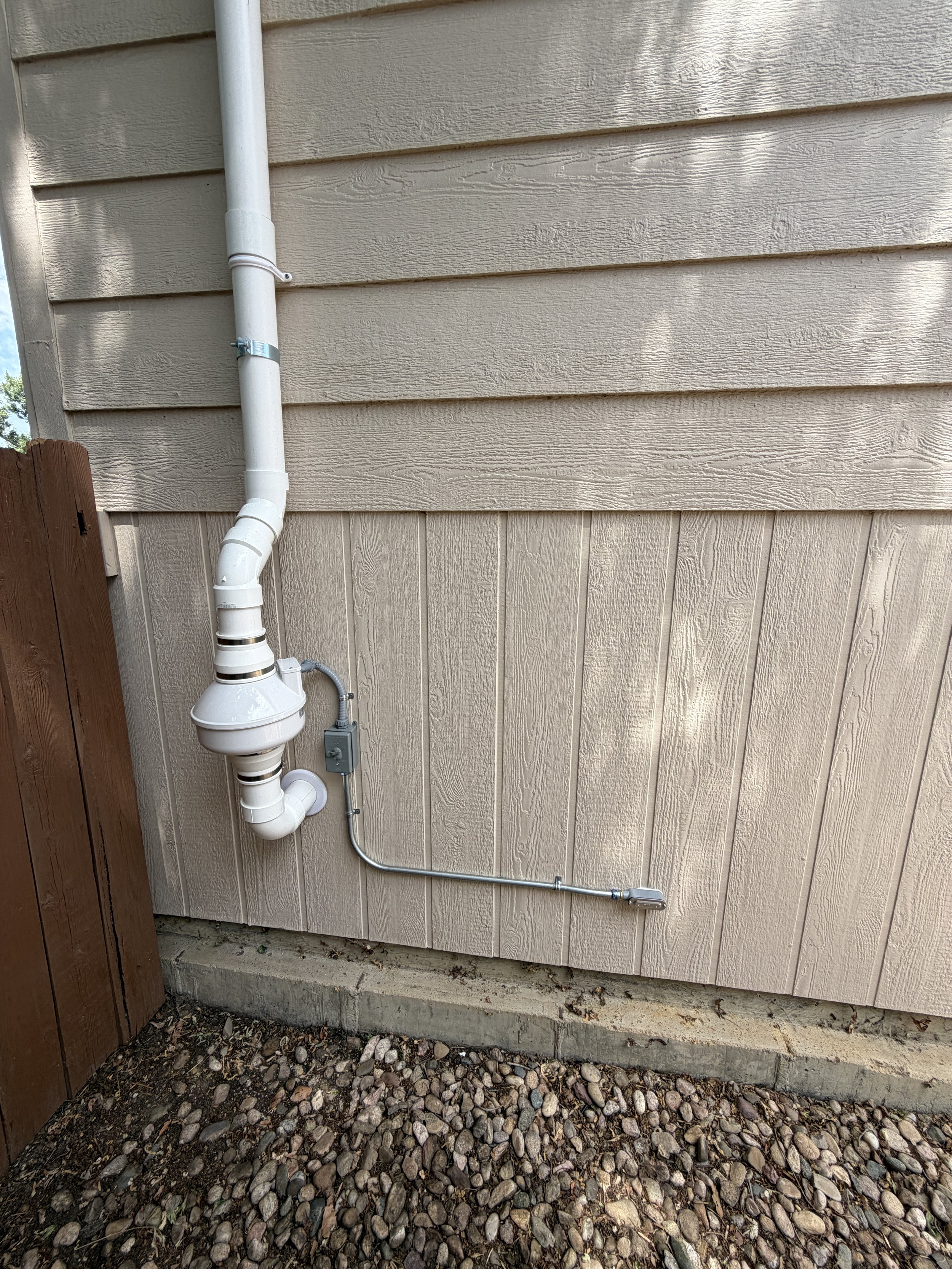 Outside view of a beige house wall with vinyl siding, a white drainage pipe with an elbow joint, and a small electrical box. The ground is covered with small rocks and dirt.