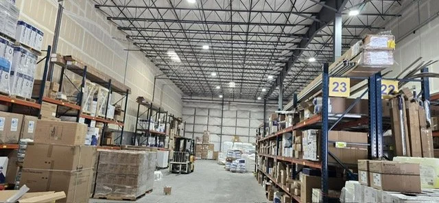 Interior view of a large warehouse with shelves stocked with various boxes and pallets, high ceiling with metal beams, and wide aisleways.