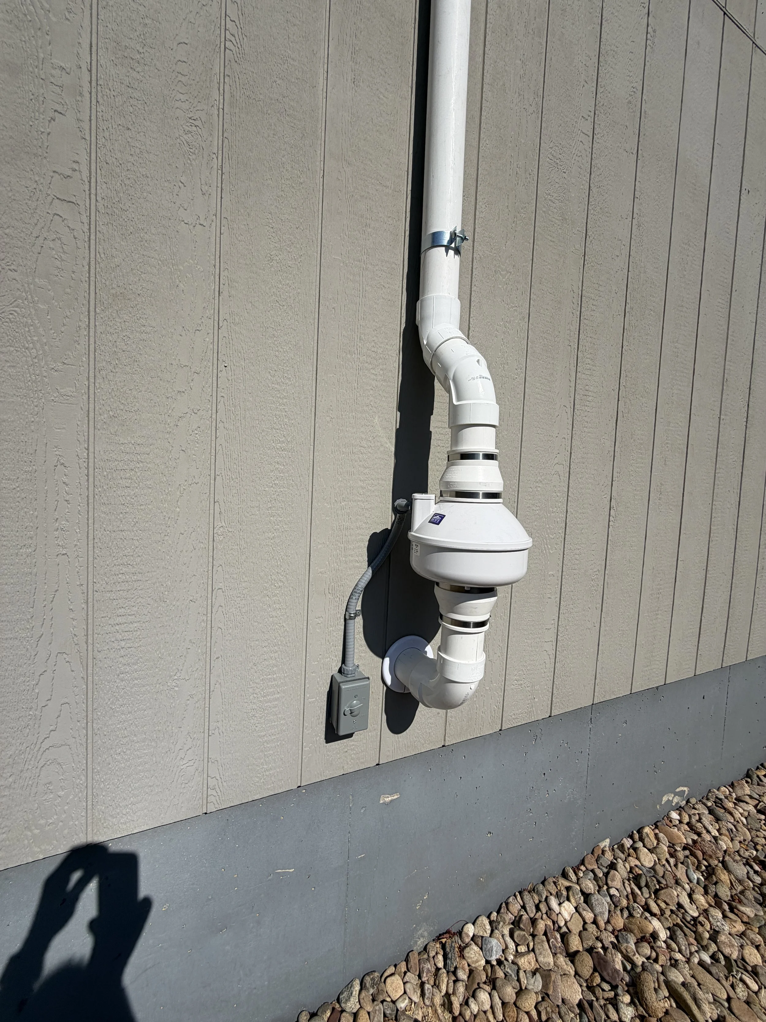 A white PVC vent pipe running vertically along the beige exterior wall of a building, secured with a metal clamp, with a small electrical box and conduit attached, and a border of small rocks at the base.