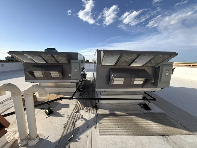 Two rooftop HVAC units installed on a flat white roof with a blue sky and clouds in the background.