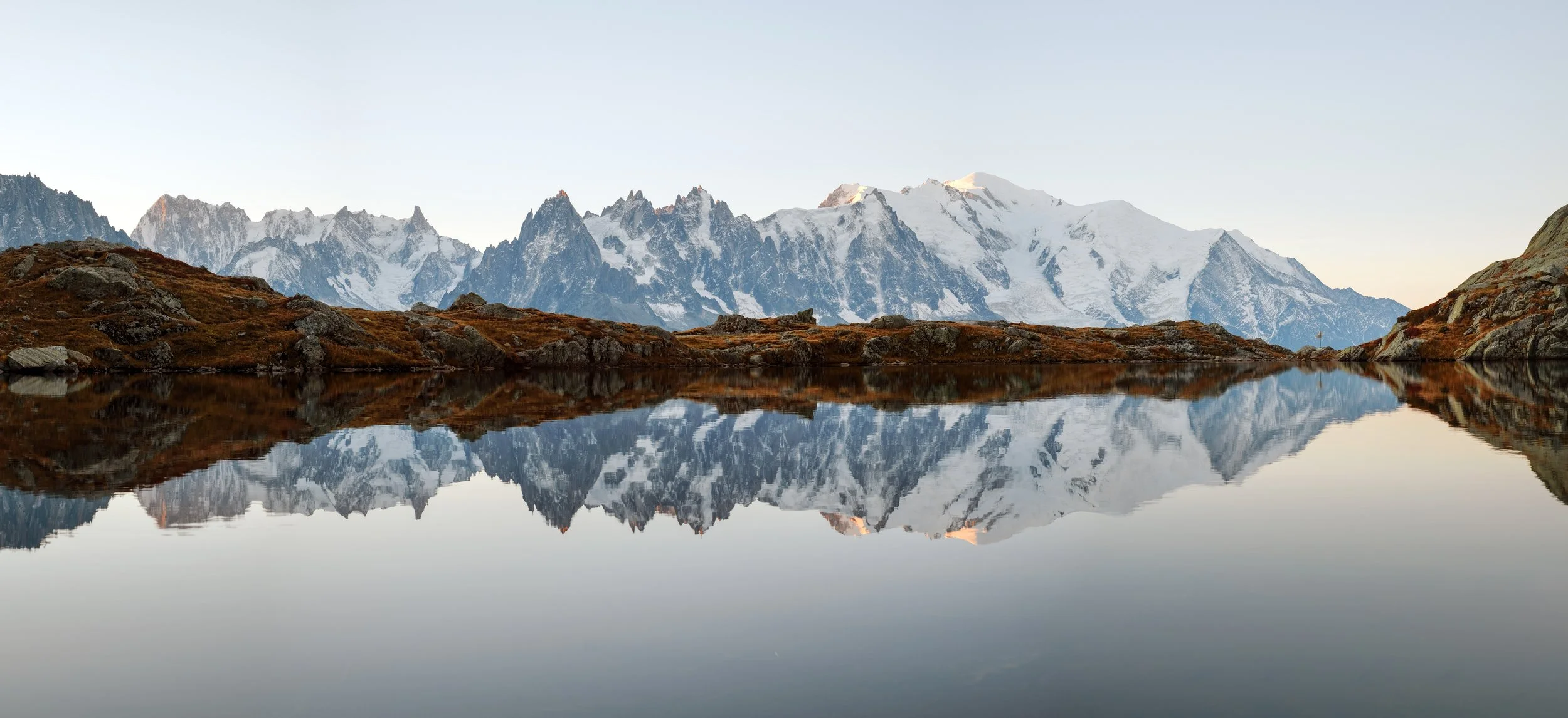 Snow-capped mountains reflecting in a calm lake with rocky terrain in the foreground.