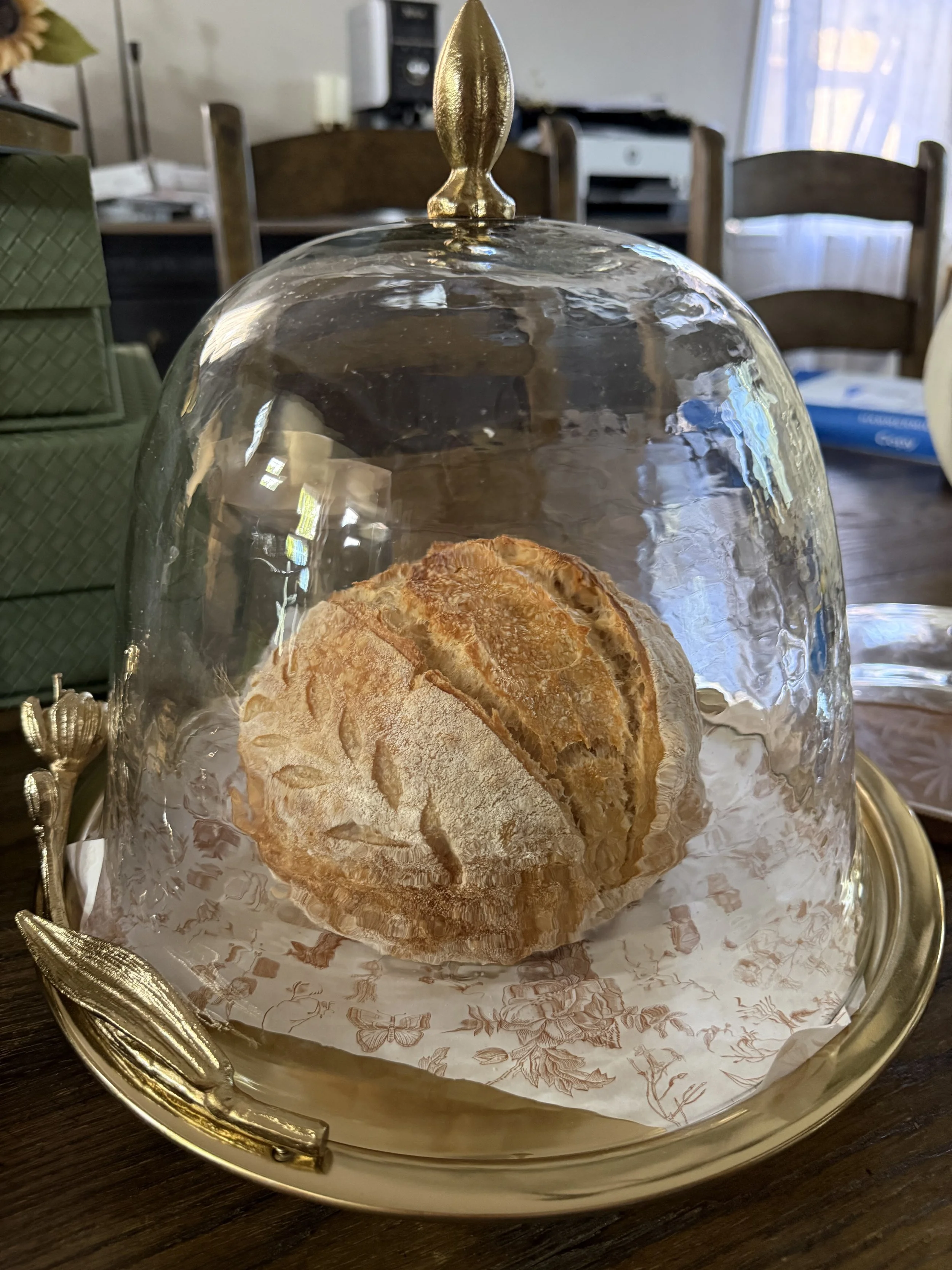 A glass cloche covering a loaf of bread on a decorative gold tray on a wooden table