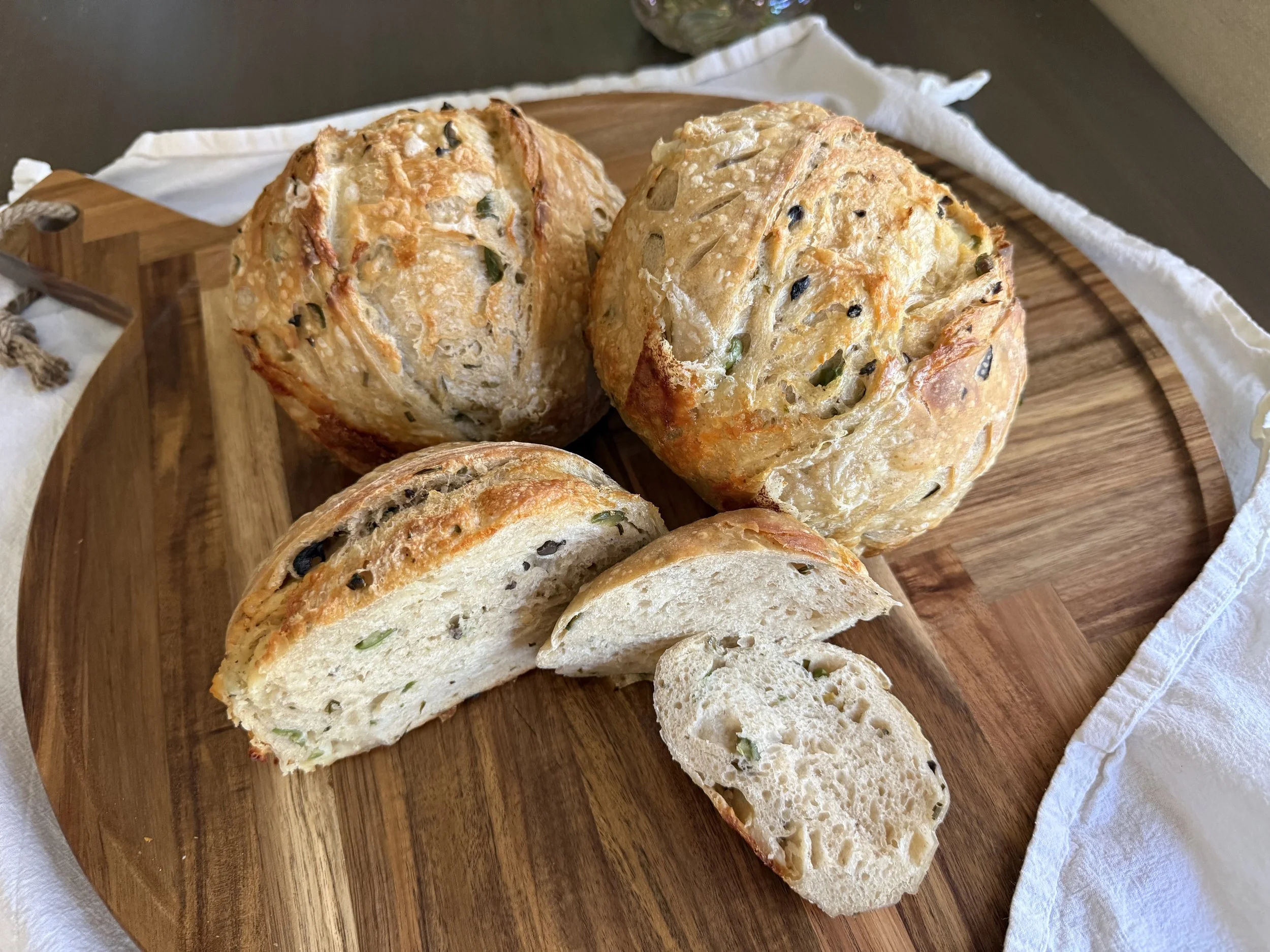 Close-up of a loaf of homemade bread with a golden crust, sliced to reveal a soft, airy interior with green herbs and black seeds, placed on a round wooden cutting board.