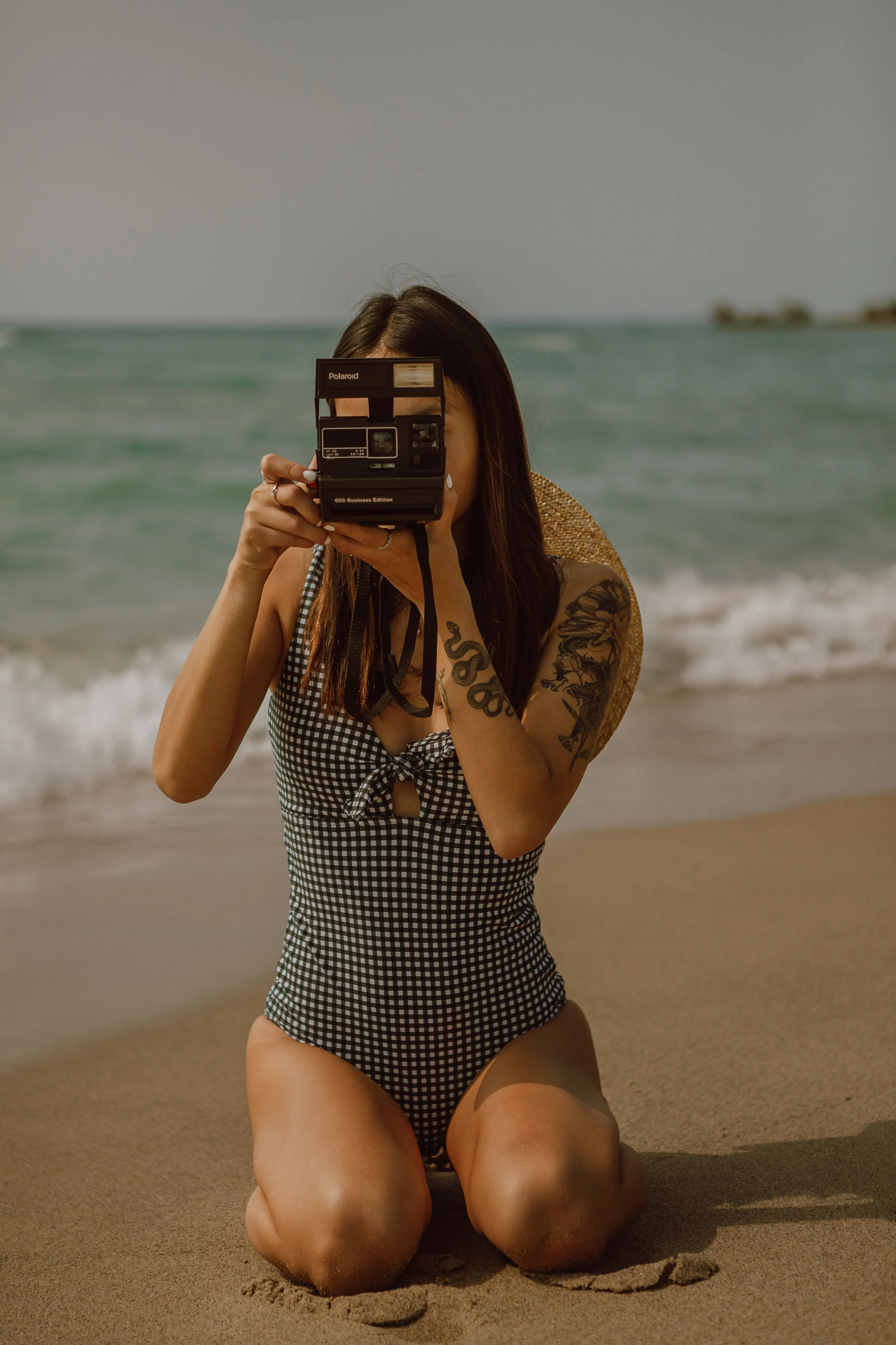 A woman with long dark hair taking a photograph with a Polaroid camera at the beach, kneeling on the sand, wearing a black and white checkered swimsuit, and holding a wide-brimmed straw hat, with the ocean and a distant shoreline in the background.
