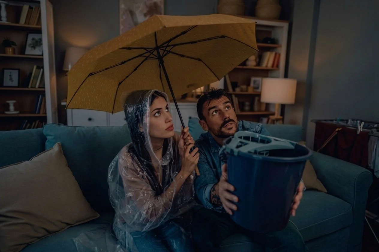A couple sitting on the couch under a yellow umbrella, catching water from a severe roof leak in a bucket.