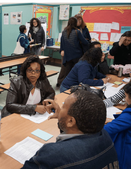 People sitting at tables and standing in a classroom, engaging in conversation and working on papers.