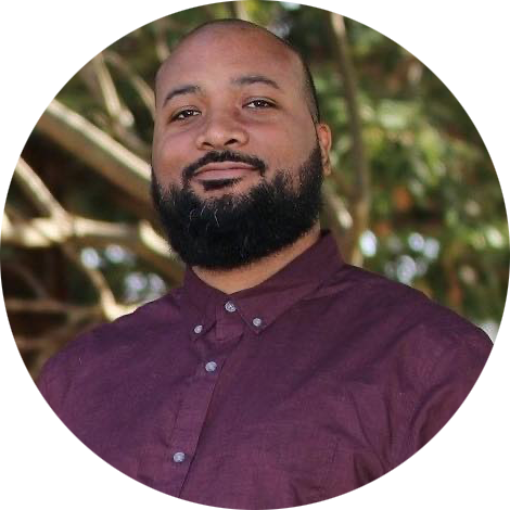 A man with a beard and mustache, wearing a maroon button-up shirt, standing outdoors near trees.