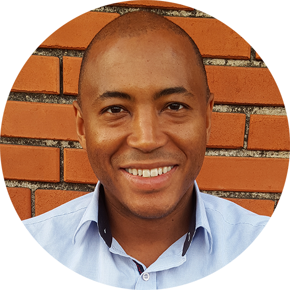 A man with a shaved head and medium skin tone smiling, wearing a light blue button-up shirt, standing in front of a brick wall.