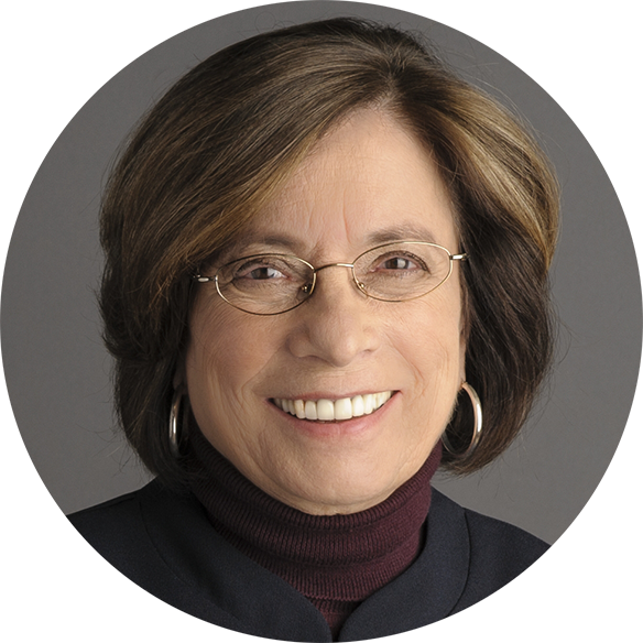 Portrait of a middle-aged woman with short brown hair, glasses, and hoop earrings, smiling at the camera against a gray background.