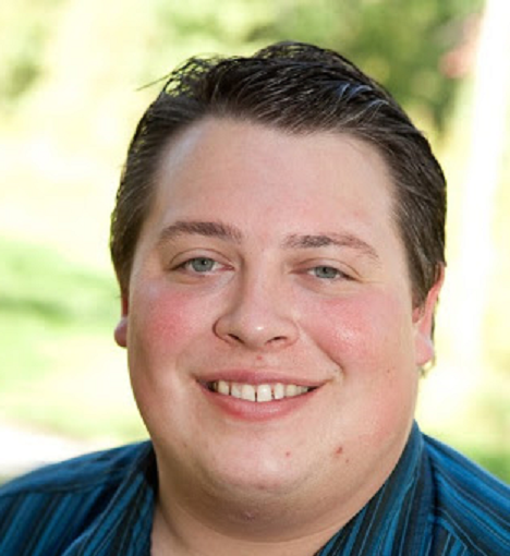 A young man with short dark hair smiling outdoors with greenery in the background.