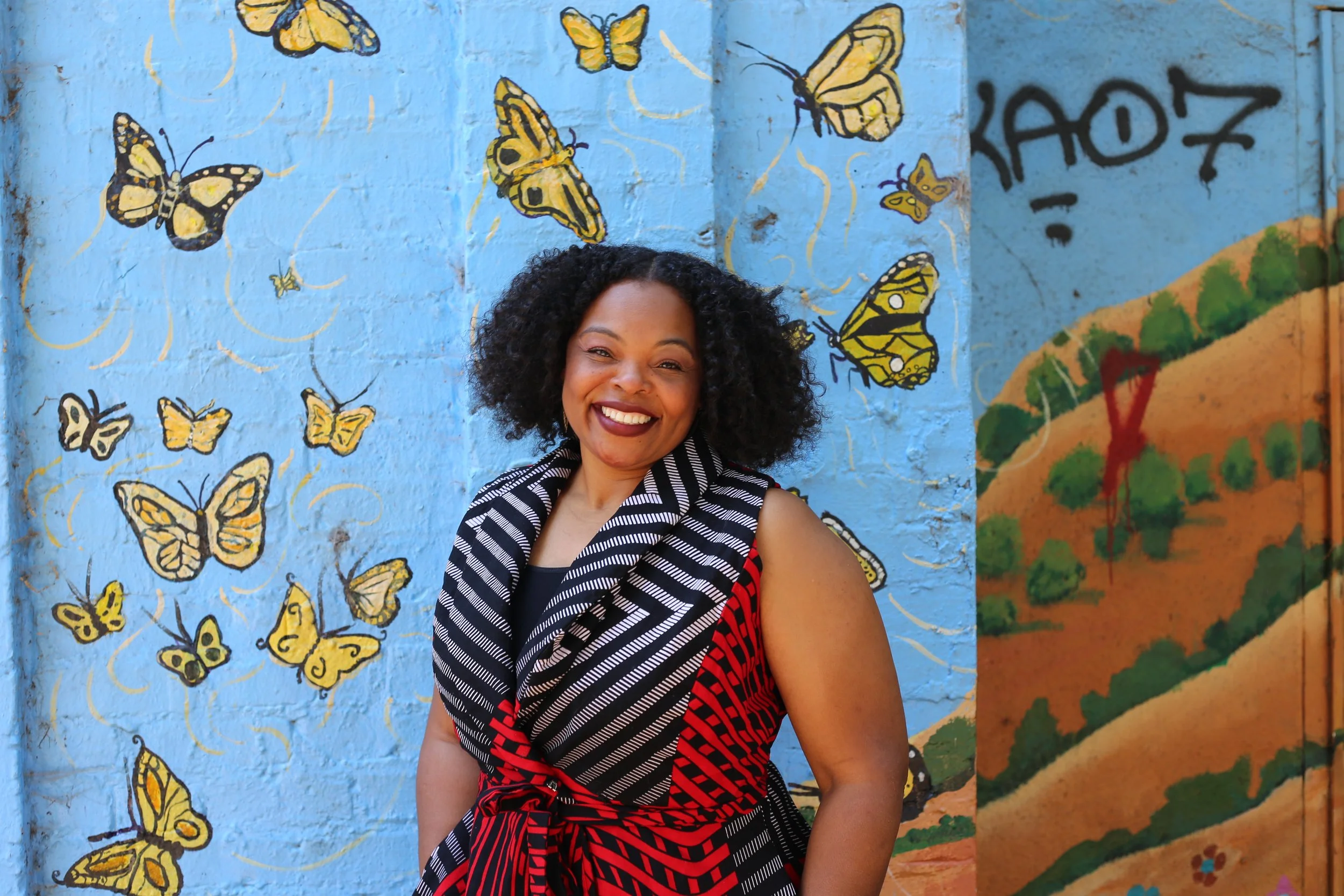 Laura, smiling, with curly black hair standing in front of a blue wall decorated with yellow butterfly murals.