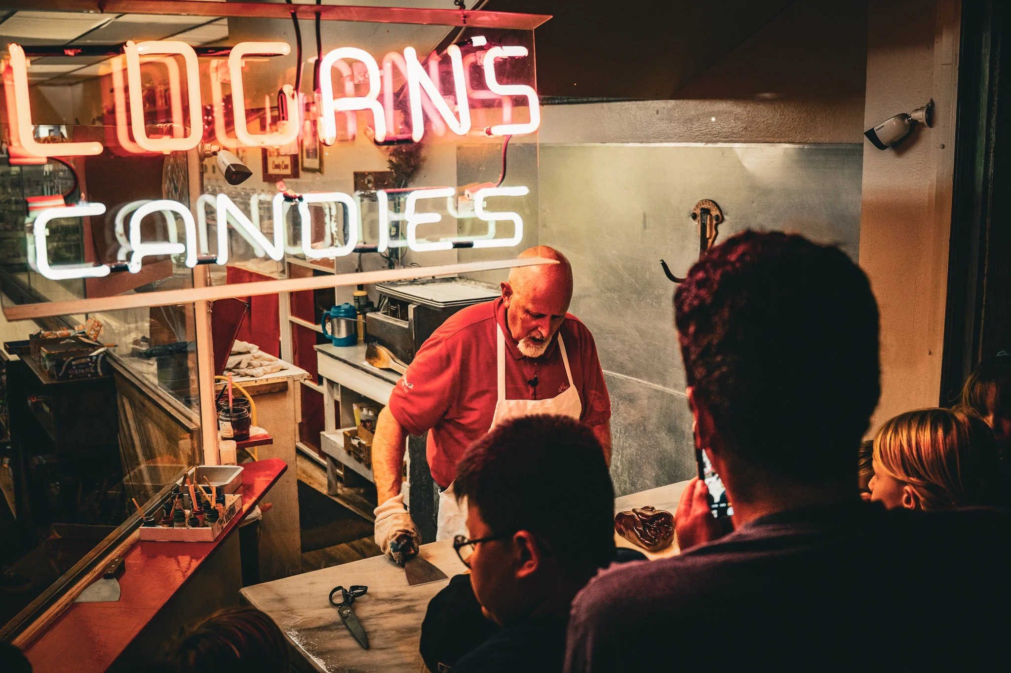 A group of children gathered around a counter inside a small shop. Logan is behind the counter working, and a neon sign inside the shop reads 'LOGANS CANDIES' in pink and white letters.