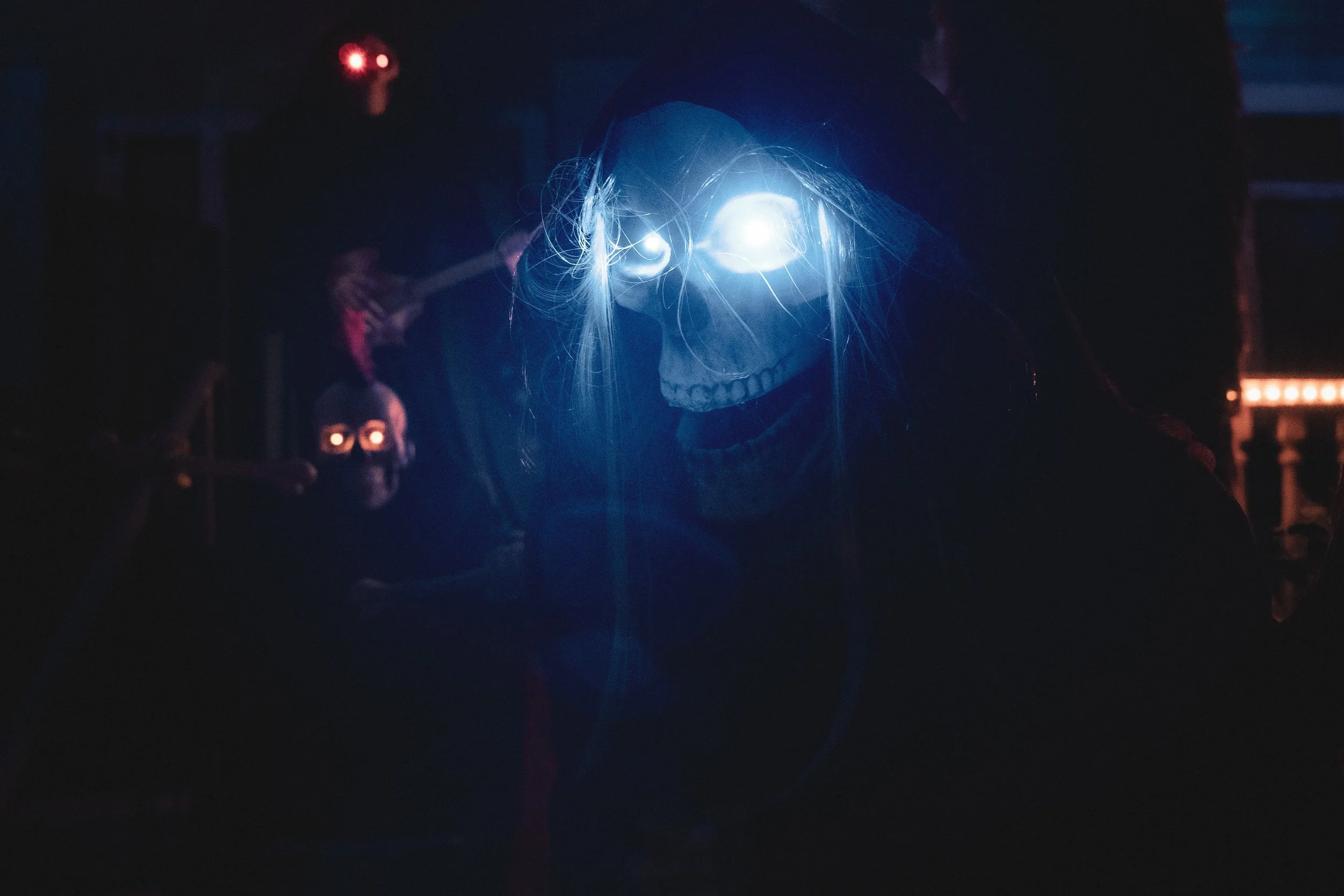 A person wearing a skeleton mask with glowing eye sockets, illuminated by blue light, in a dark environment with other skeleton masks in the background.