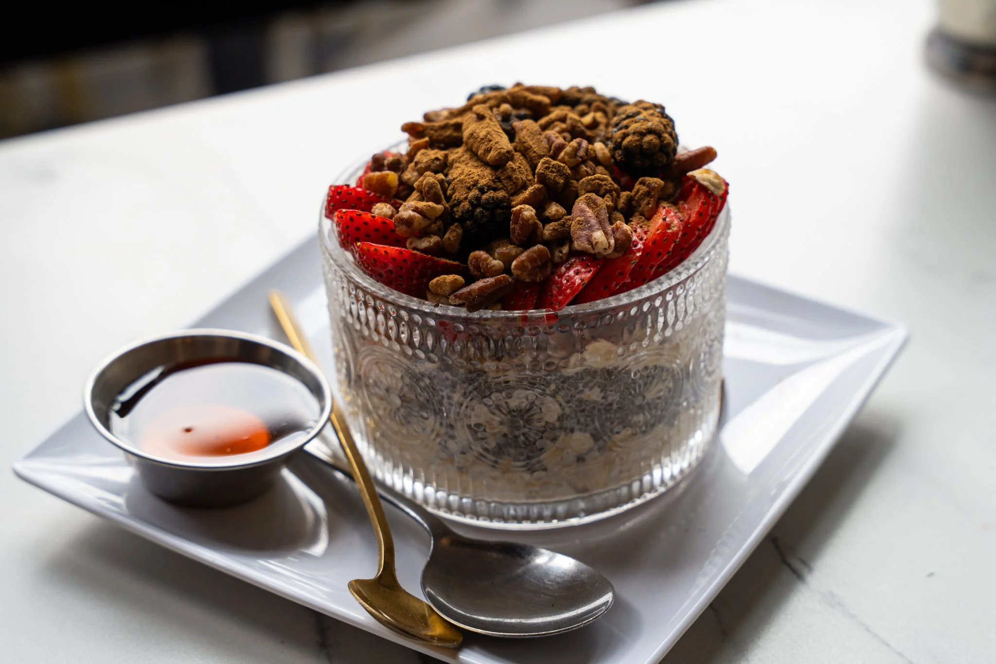 A dessert in a glass bowl with strawberries and chocolate chips on top, along with a small glass pitcher of syrup and a gold spoon on a white tray.