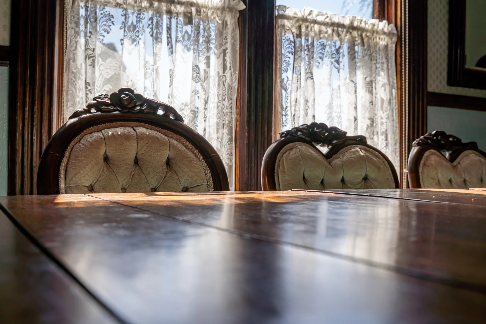 Vintage wooden dining table with carved backrest chairs in front of lace curtains on windows.