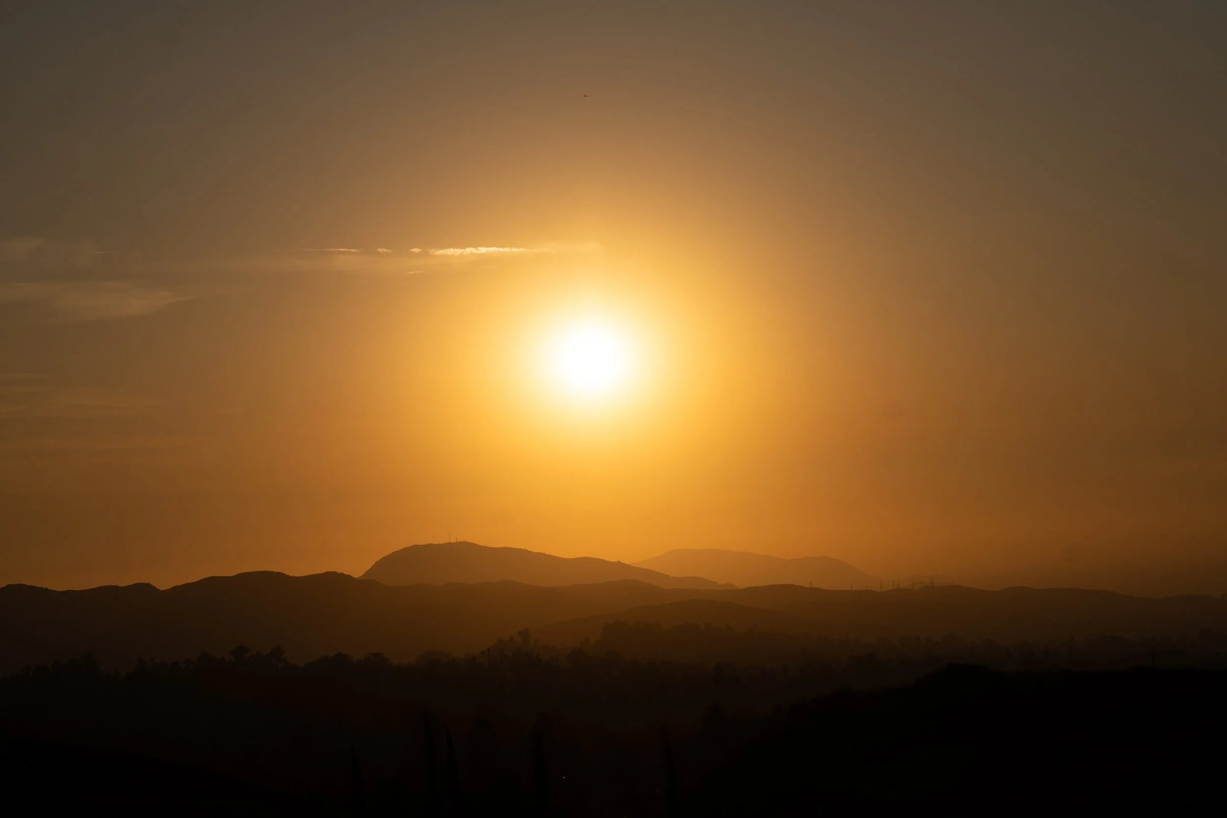 Sunset over mountains with a bright sun low on the horizon and dark silhouette of hills in the foreground.