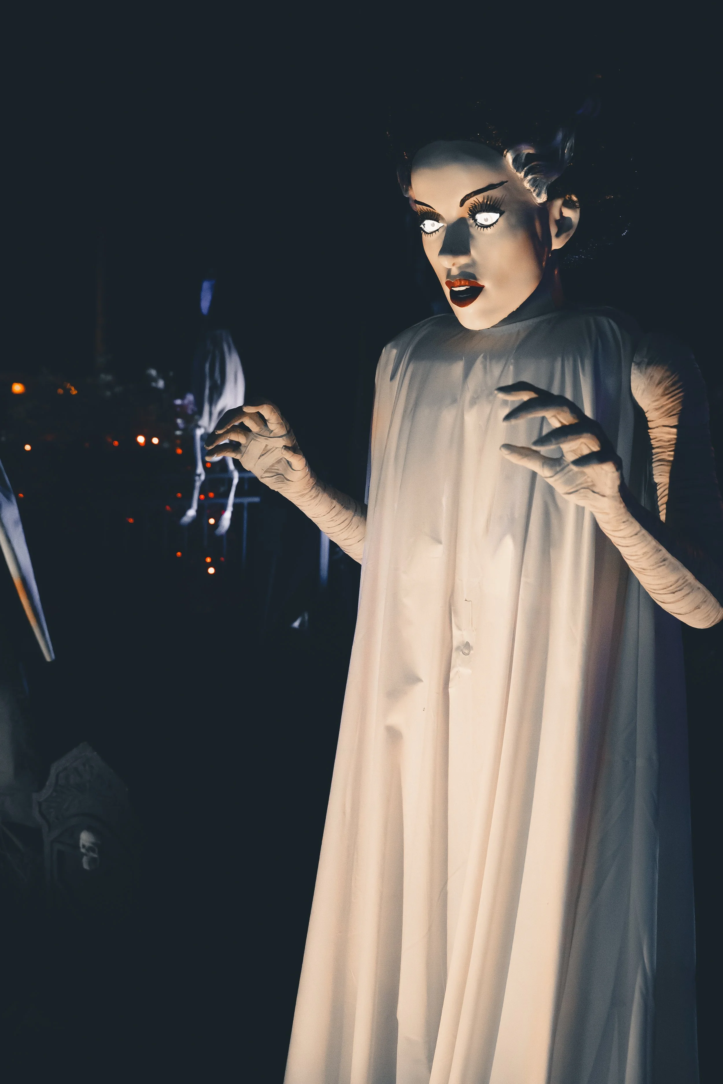 A person wearing a large, eerie mask with glowing white eyes, dark makeup, red lipstick, and a flowing white gown with long gloves, standing in low lighting, creating a spooky atmosphere.