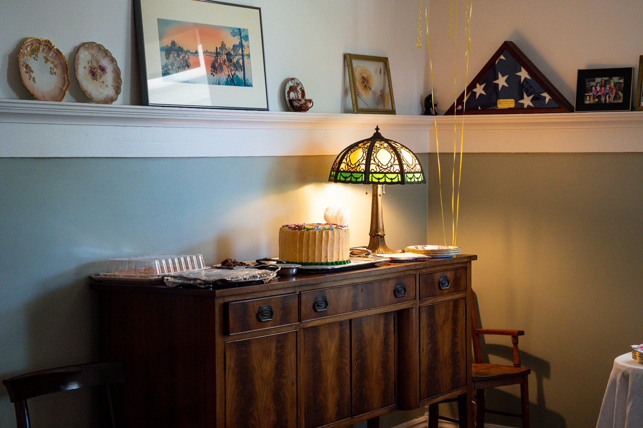 A wooden sideboard with a large, round cake decorated with colorful sprinkles on top, surrounded by serving utensils. A Tiffany-style lamp with a stained glass shade is lit and positioned on the sideboard. On the wall above, decorative plates, framed