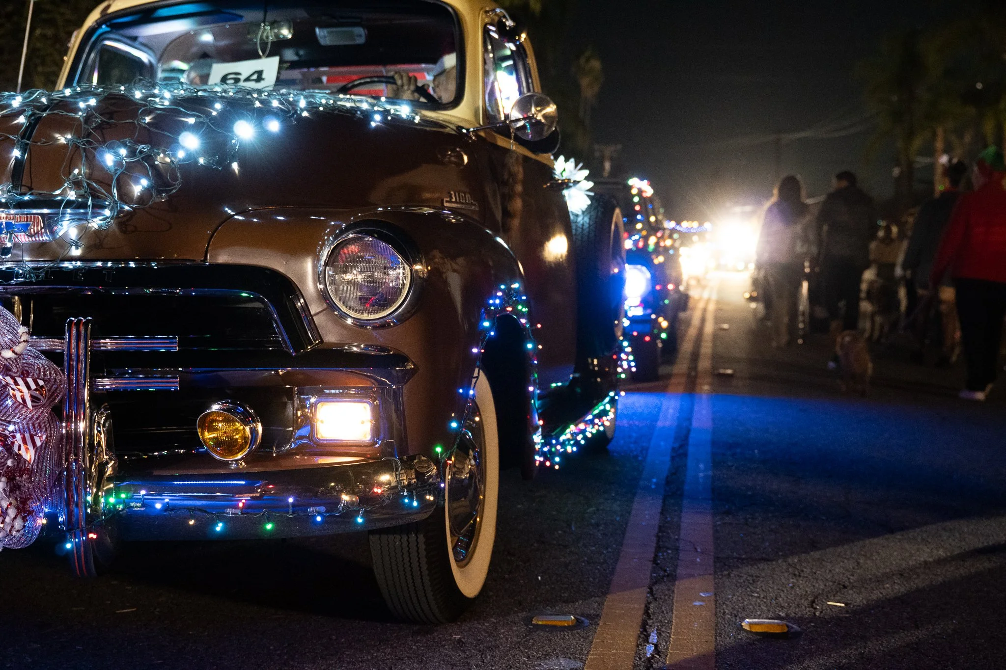 A vintage brown car decorated with white and multicolored Christmas lights parked on a street at night during a parade or celebration, with people in the background.