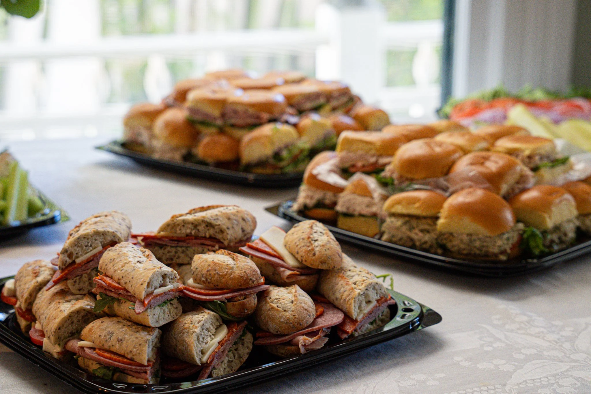 Tray of assorted sandwiches, including sub sandwiches and roll-ups, on a table in front of a window.