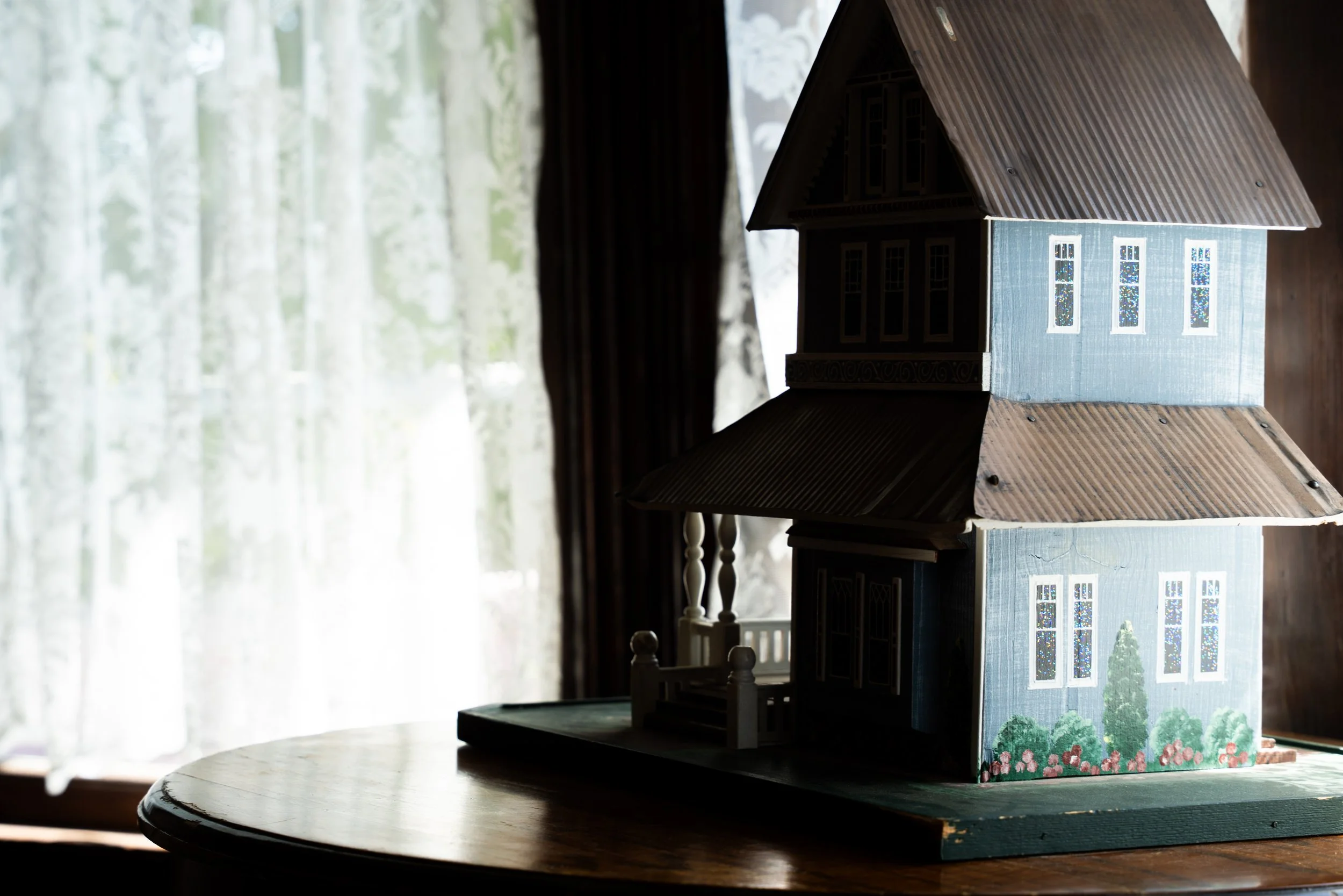 A miniature blue house with white trim and four windows, sitting on a dark wooden table near a window with sheer curtains.
