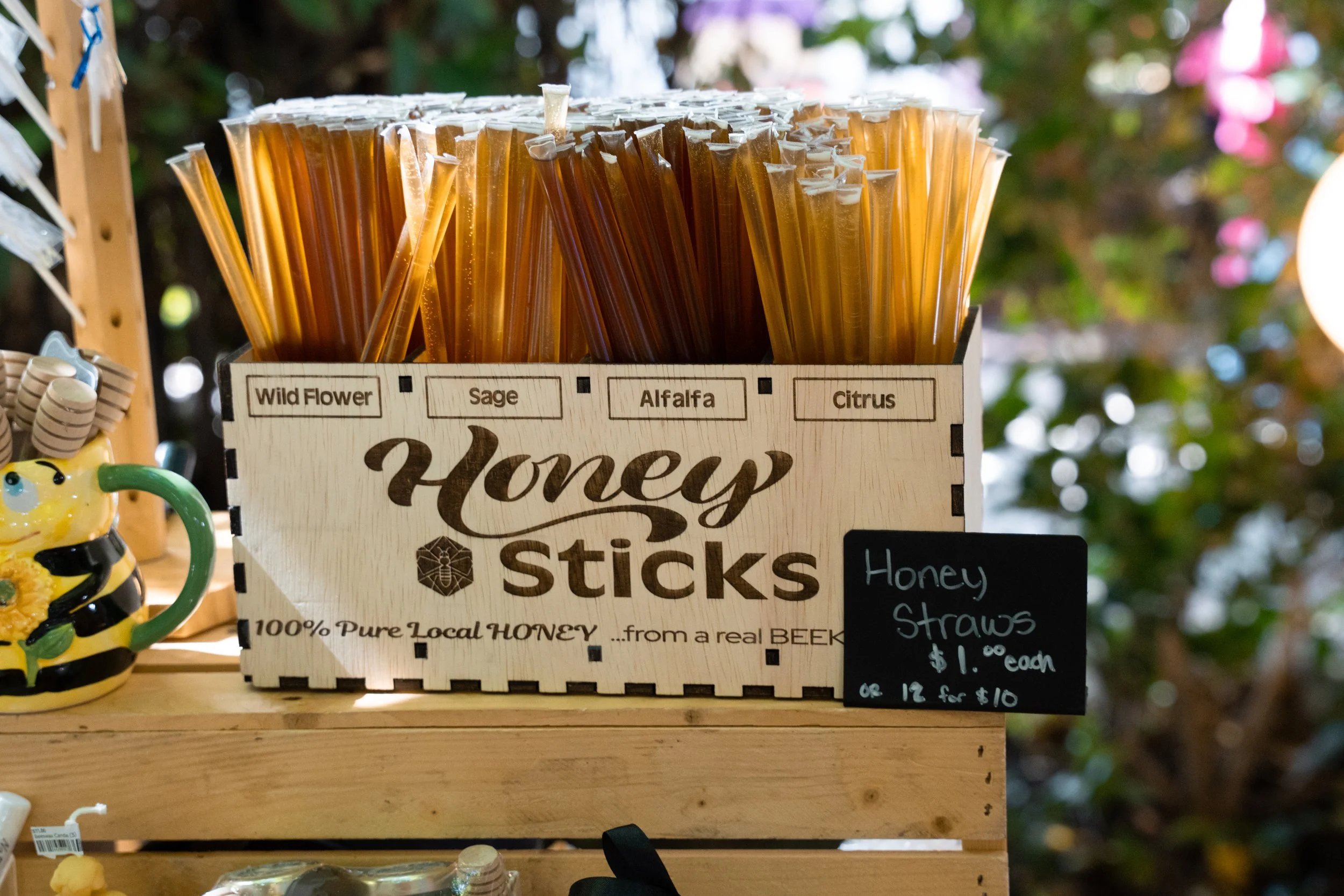 Plastic honey sticks in a wooden display box, labeled with flavors including Wild Flower, Sage, Alfalfa, and Citrus. A small chalkboard sign indicates honey straws are $1.00 each or 12 for $10.00. A ceramic bee and honey pot are also visible in the d