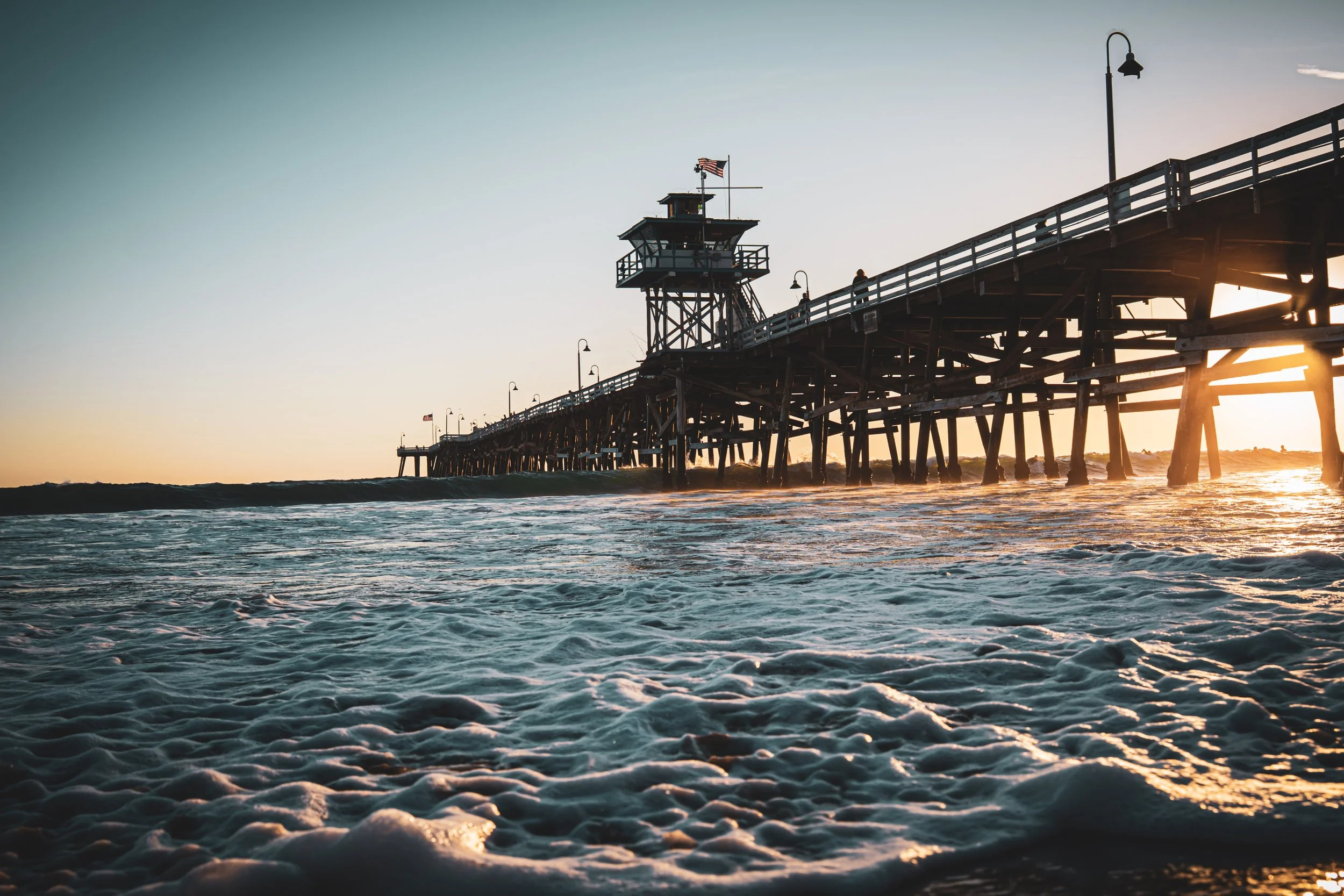 A wooden pier extending into the ocean during sunset, with a lighthouse and American flags on top, and some people walking along the pier, with waves crashing in the foreground.