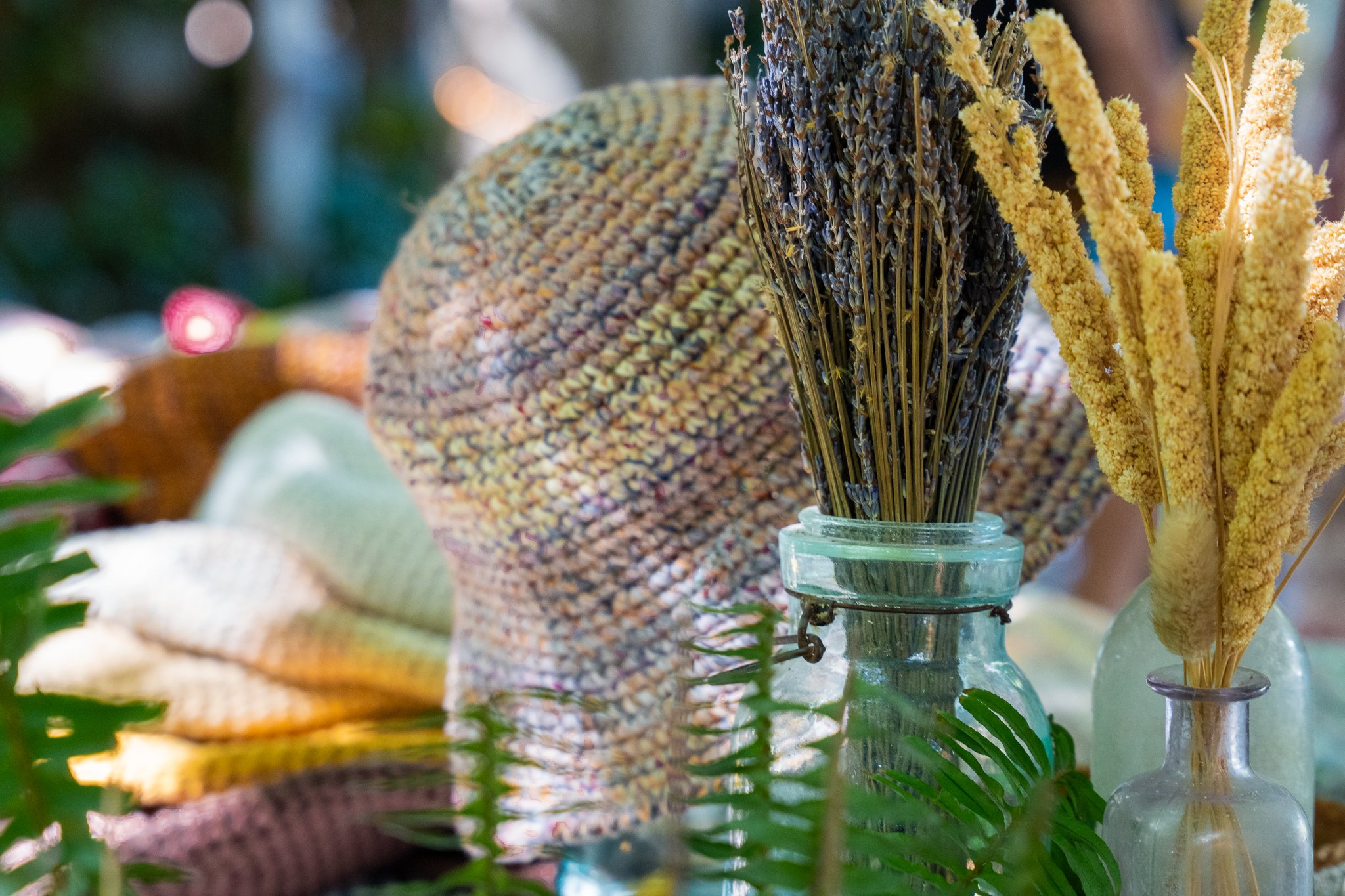 Close-up of a variety of dried flowers and herbs in a glass jar, with a background of blurred greenery and warm lights.