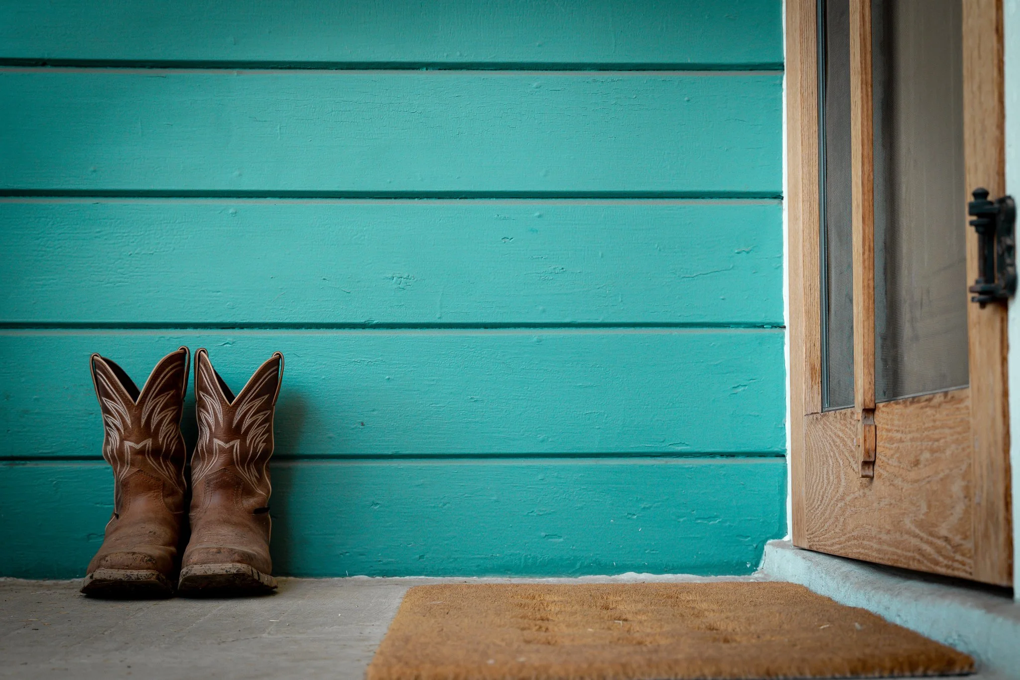 Pair of brown cowboy boots placed on a concrete floor in front of a teal wooden wall and a door with a small window and black hardware.
