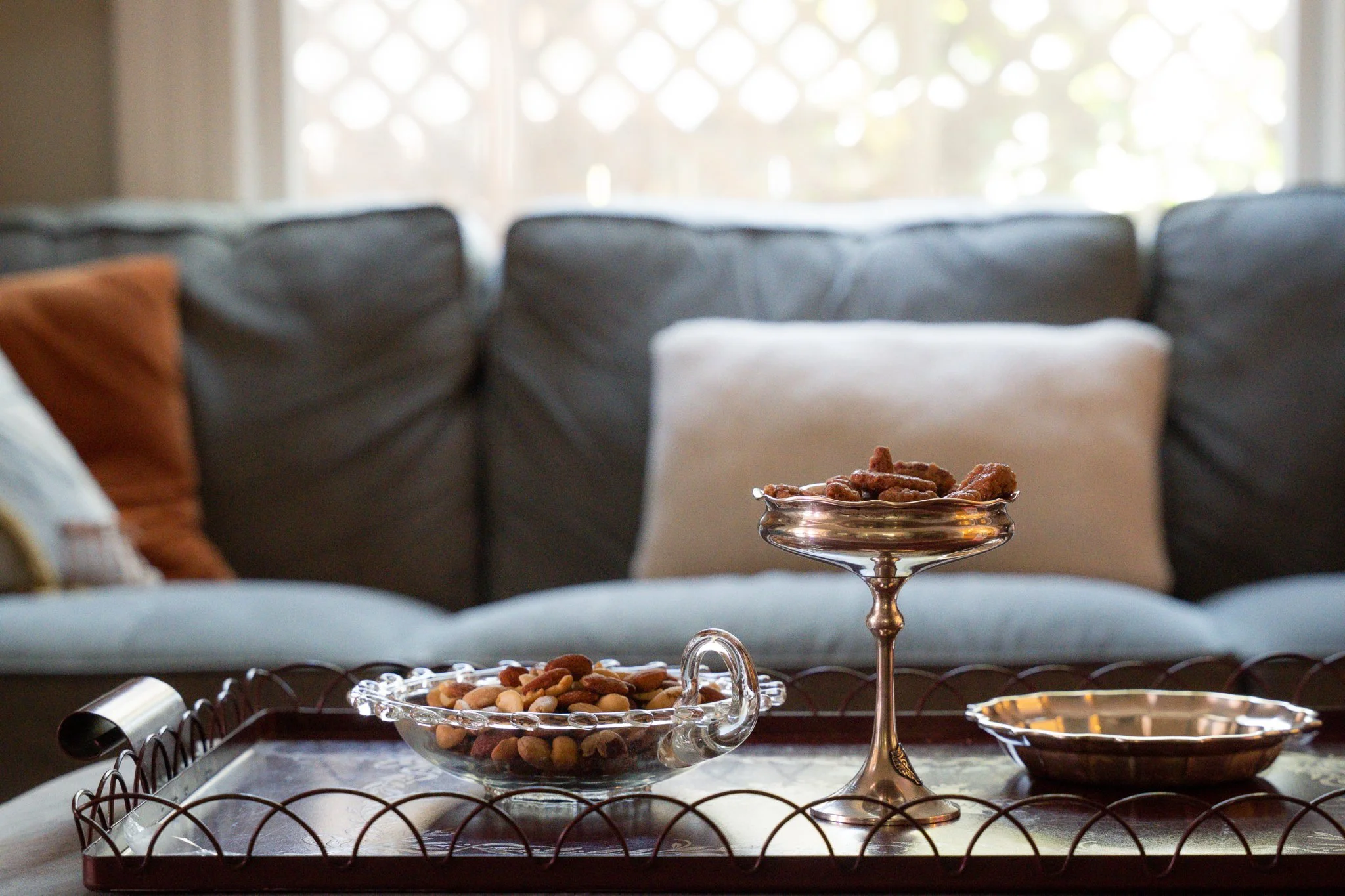 A glass bowl filled with mixed nuts sits on a tray, and a tall, ornate glass dish with more mixed nuts is behind it, on a table in front of a blurred background of a couch and a window with sunlight.