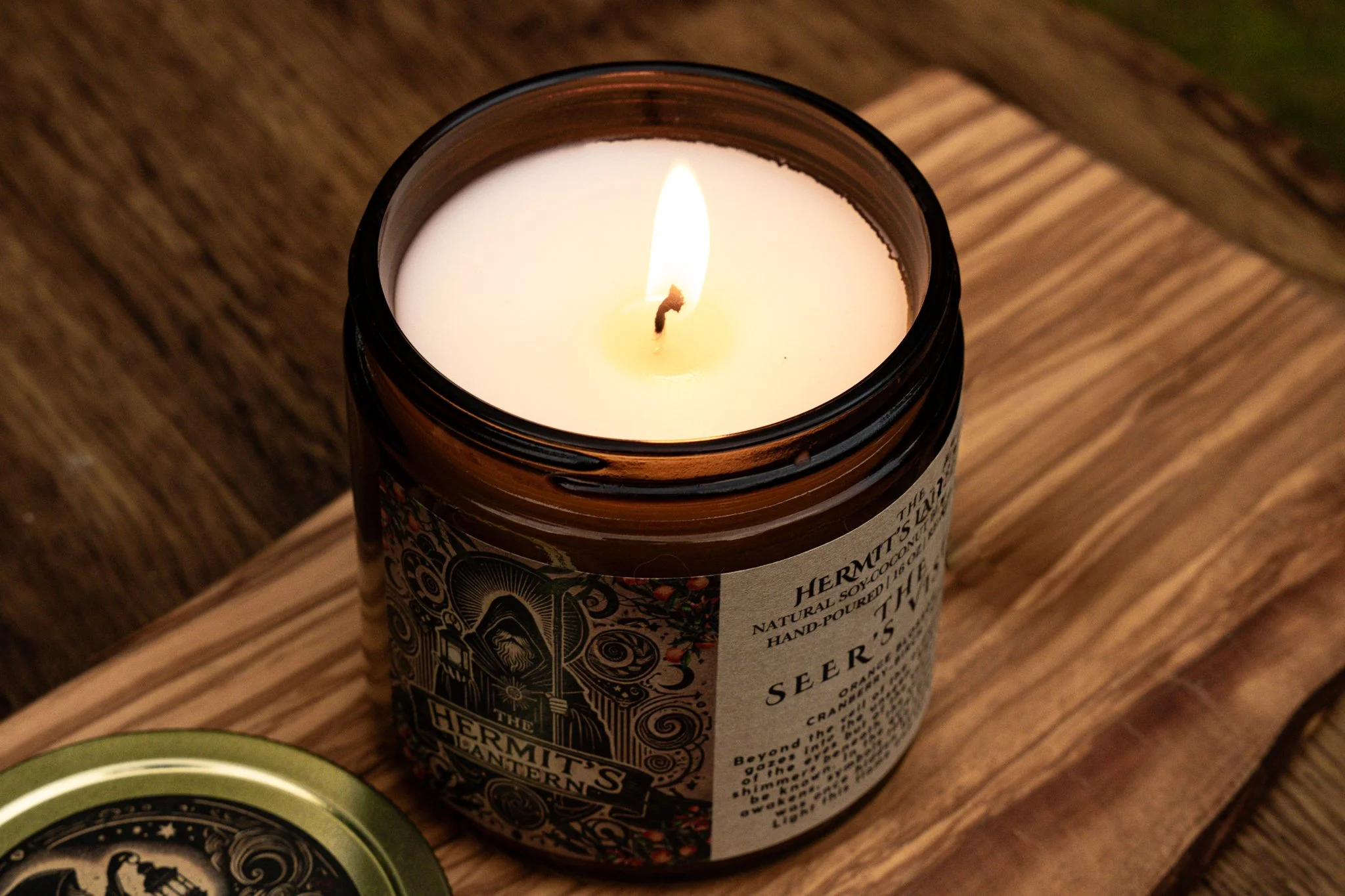A lit candle in a dark glass jar with decorative label on a wooden surface.