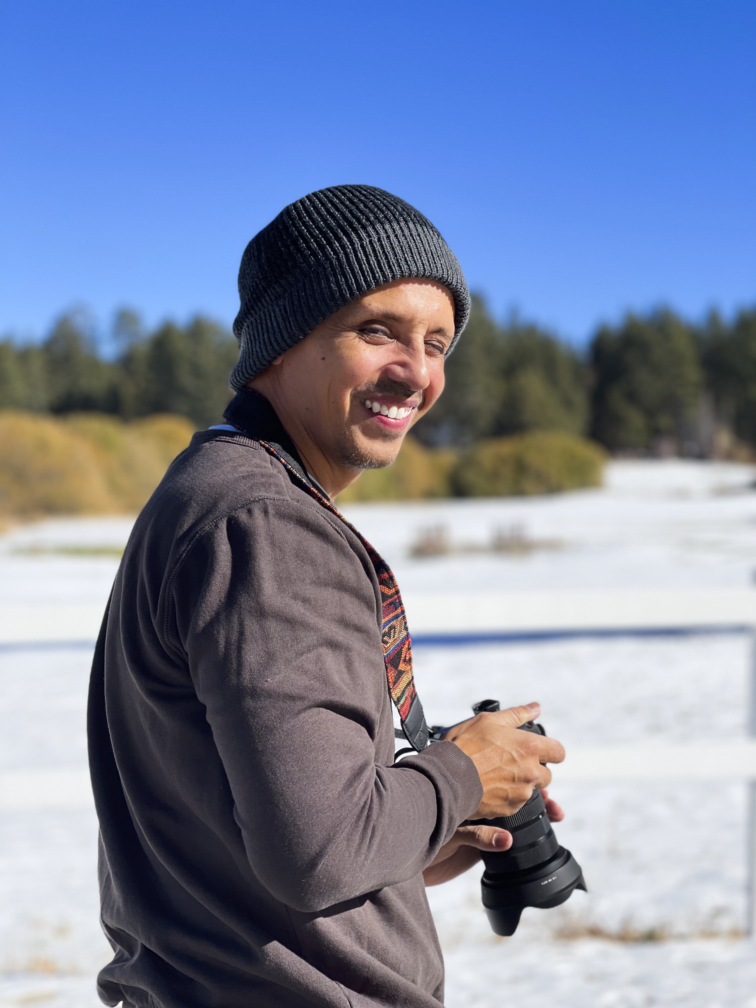 A man with a gray beanie and gray jacket holding a camera with a lens, standing outdoors on snow-covered ground with a backdrop of trees and a clear blue sky.