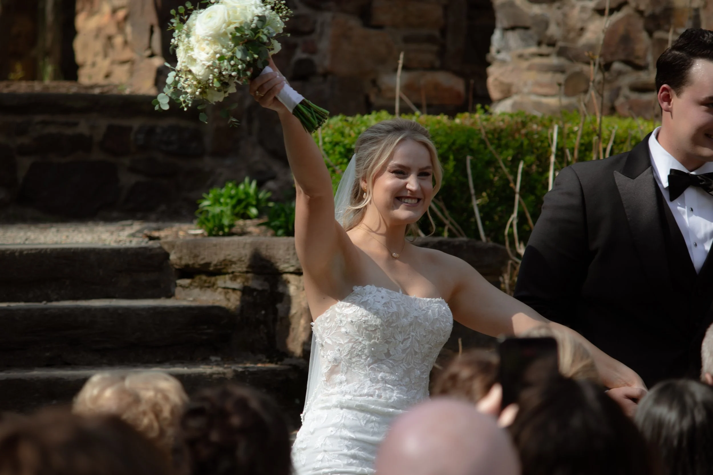 A bride in a strapless white lace wedding gown smiling and raising a bouquet of white flowers during her wedding outdoors, with a groom in a black tuxedo beside her.
